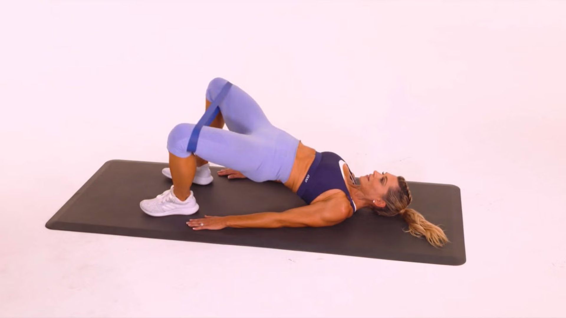 A woman performs a glute bridge exercise on a black mat, wearing a blue sports bra and leggings, a resistance band above her knees, and white sneakers.