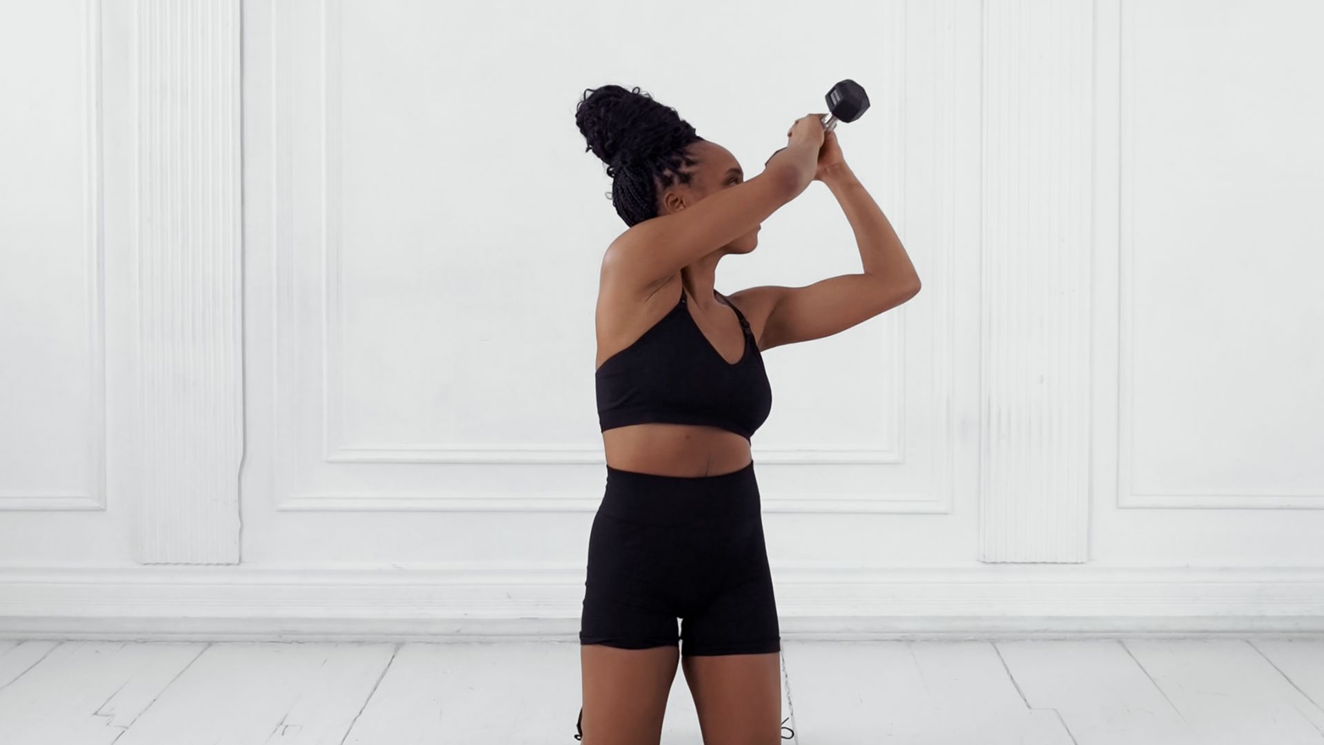Woman in black sportswear exercises indoors with a dumbbell, lifting it overhead. The room is minimalistic with white paneled walls.