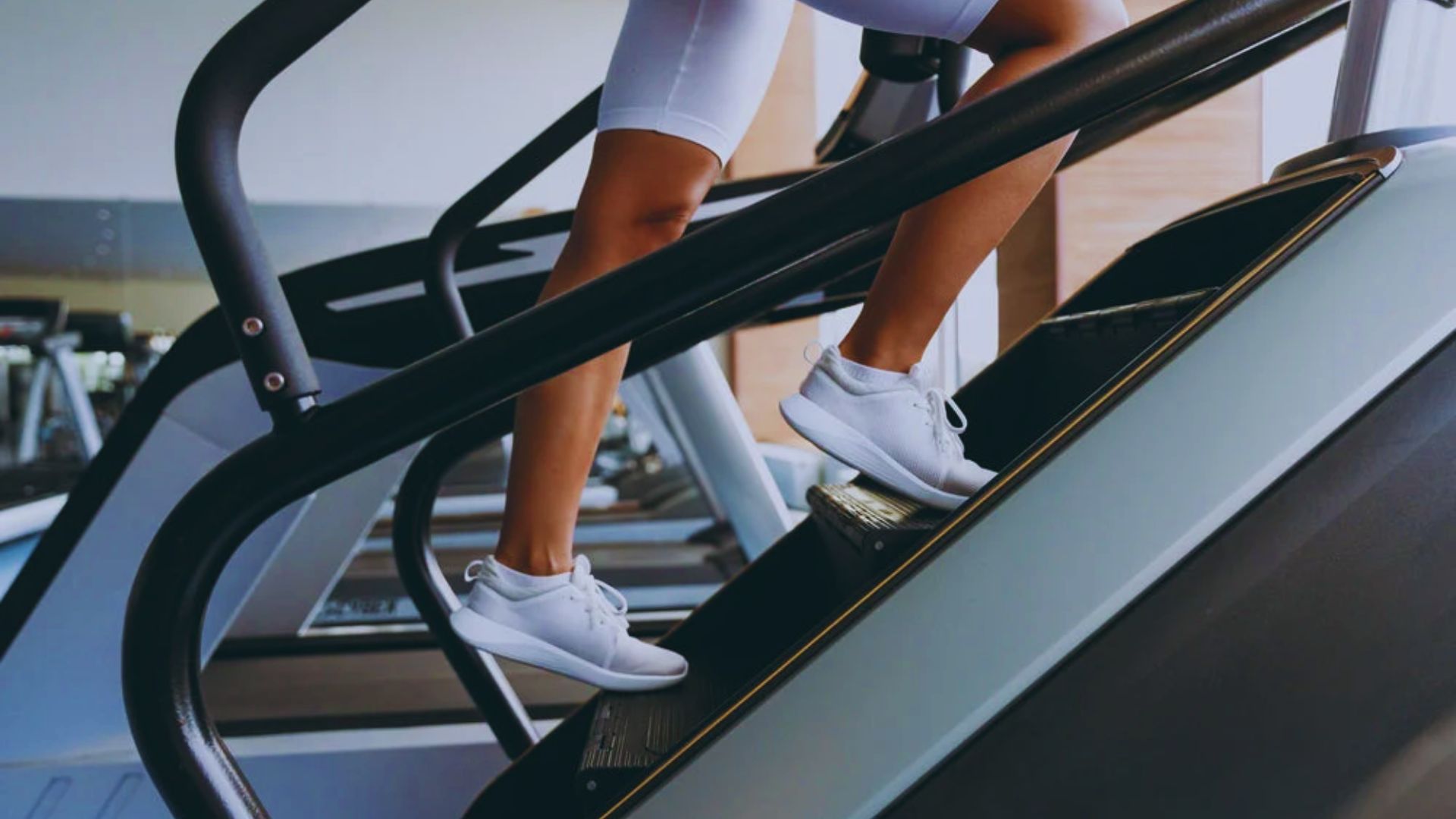 A person wearing white sneakers and shorts exercises on a stair machine in a gym. The setting is bright and modern, conveying a sense of energy and focus.