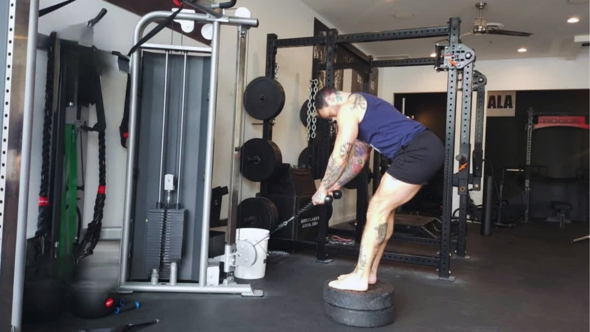 A tattooed individual in a navy tank top and black shorts exercises using a cable machine. They stand on a platform in a gym, focused and determined.