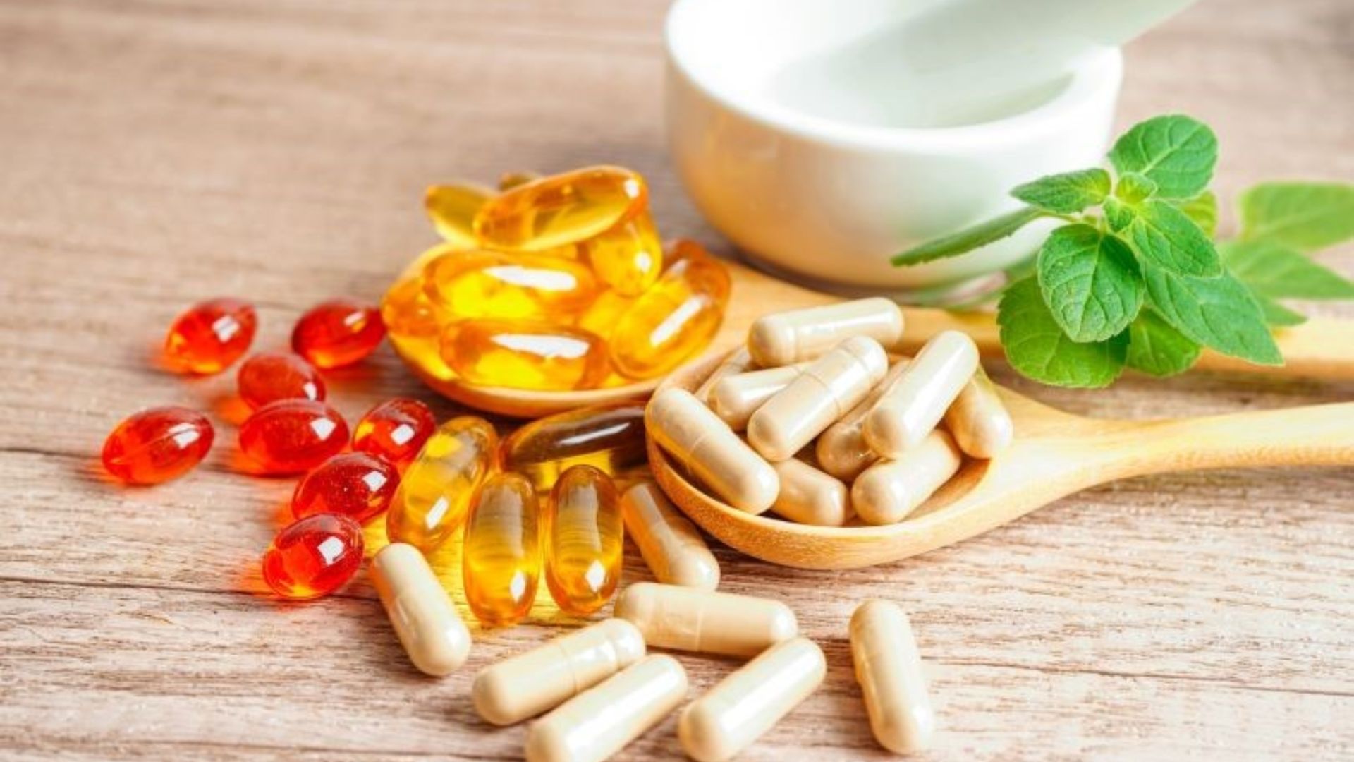 Assorted capsules and gel pills on wooden spoons with green leaves; next to them, a white mortar and pestle. The scene conveys health and wellness.