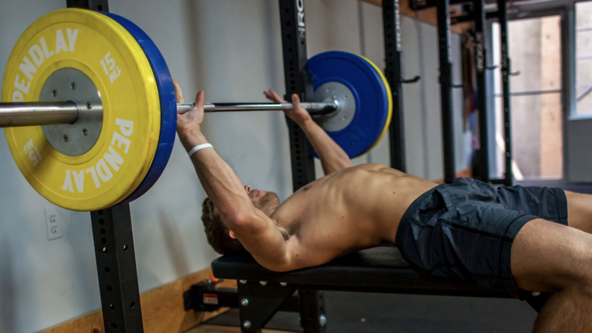 A shirtless person on a bench press lifts a barbell with blue and yellow weights in a gym, showcasing strength and focus.