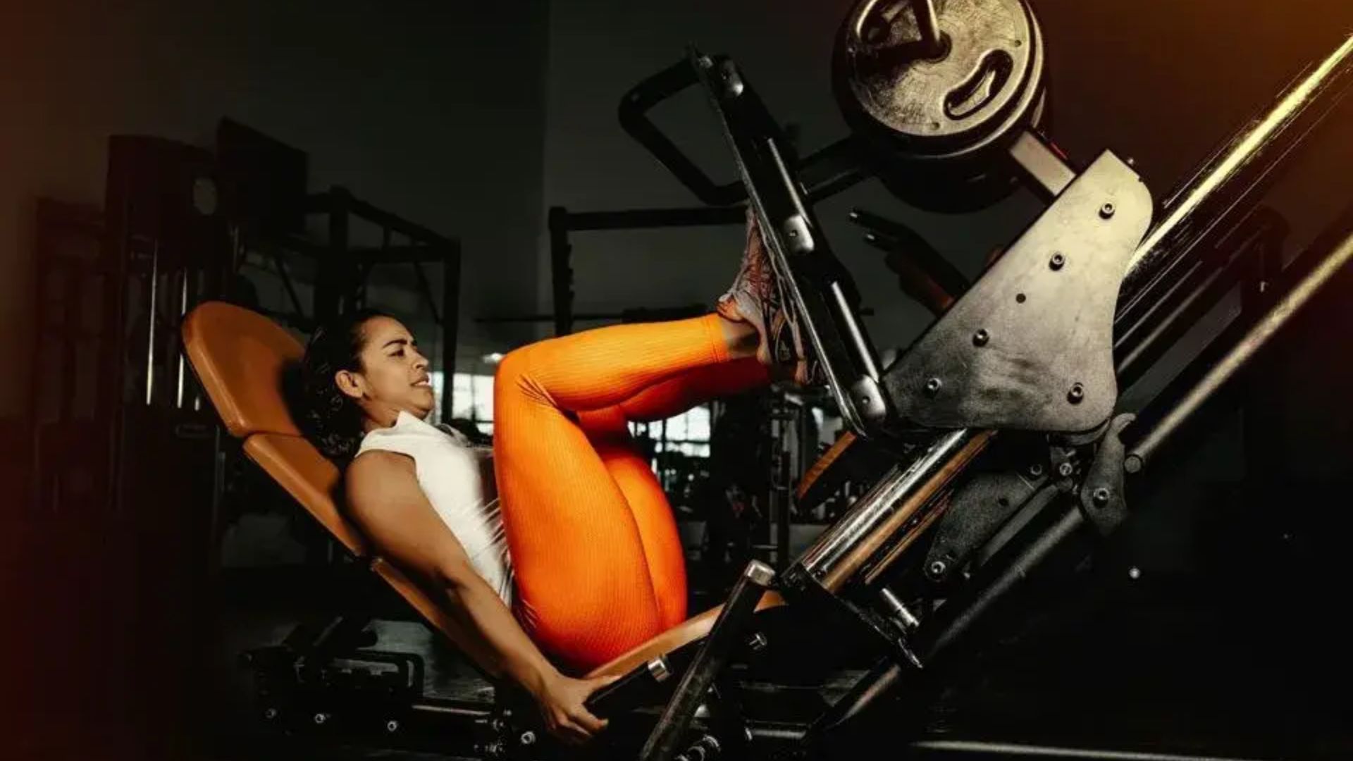 A woman in bright orange leggings uses a leg press machine in a dimly lit gym, showing focus and determination during her workout.