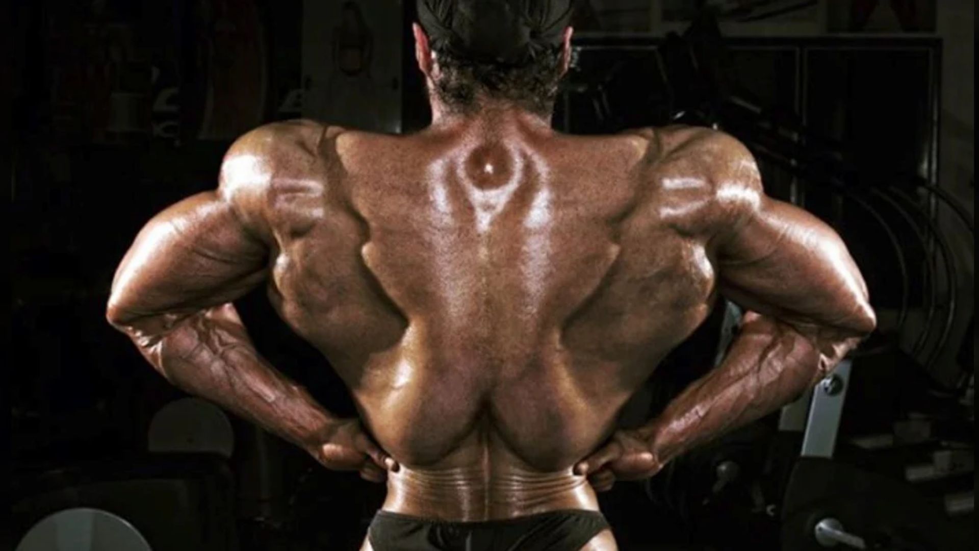 A muscular bodybuilder stands with their back facing the camera, showcasing a highly defined and muscular back and arms. The gym setting is dimly lit.