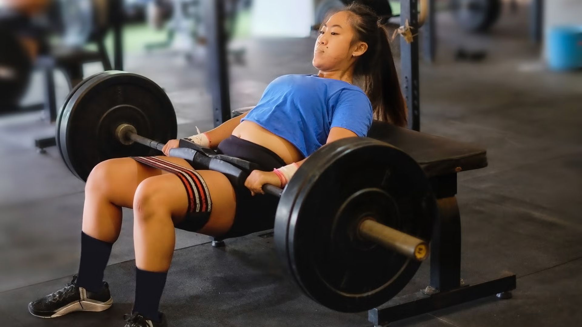 A woman sits on a bench, holding a barbell, focused on her workout in a gym setting.