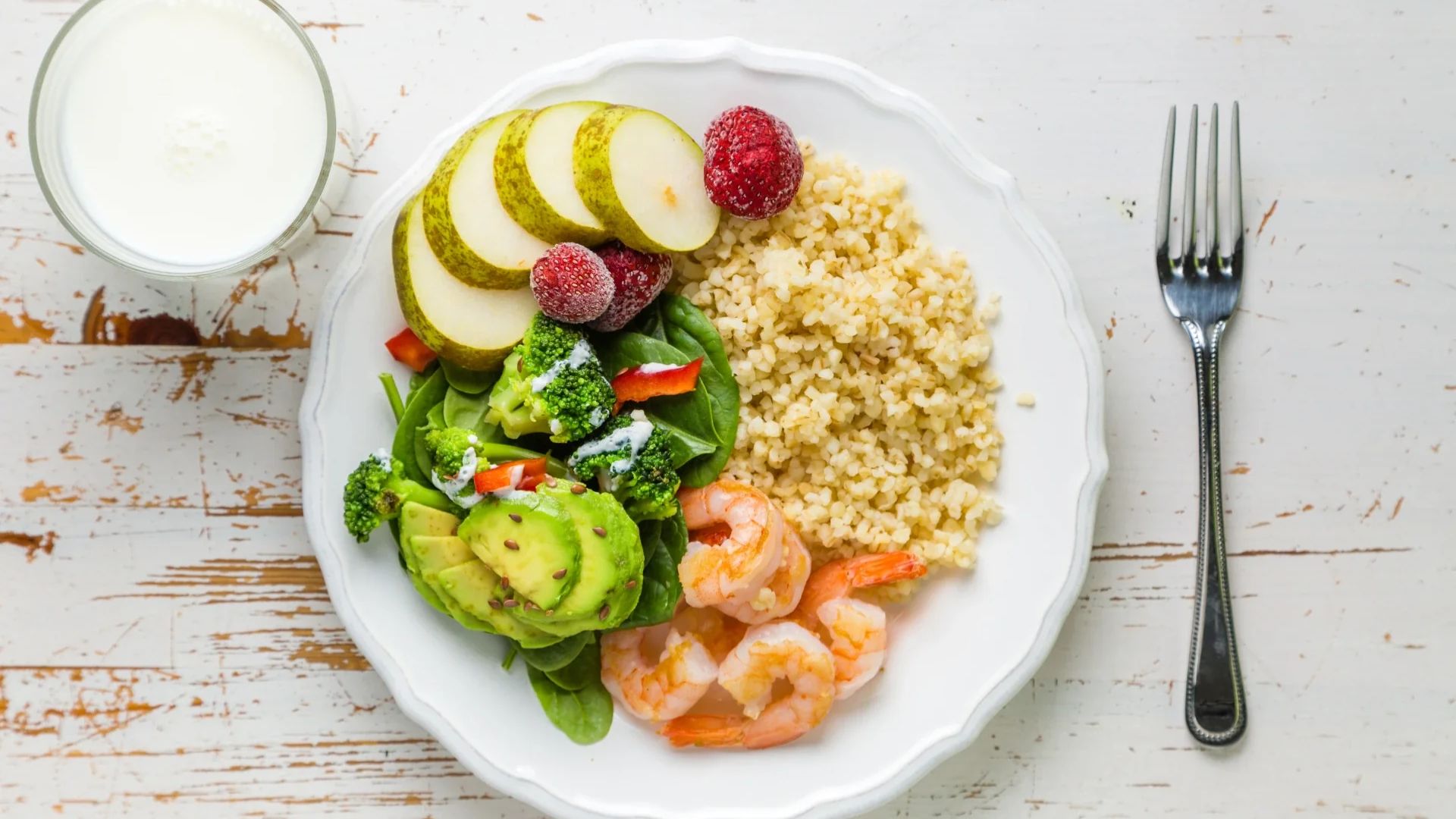 A white plate features sliced pears, raspberries, quinoa, shrimp, avocado, spinach, and broccoli, next to a fork. A glass of milk sits on a rustic wooden table.