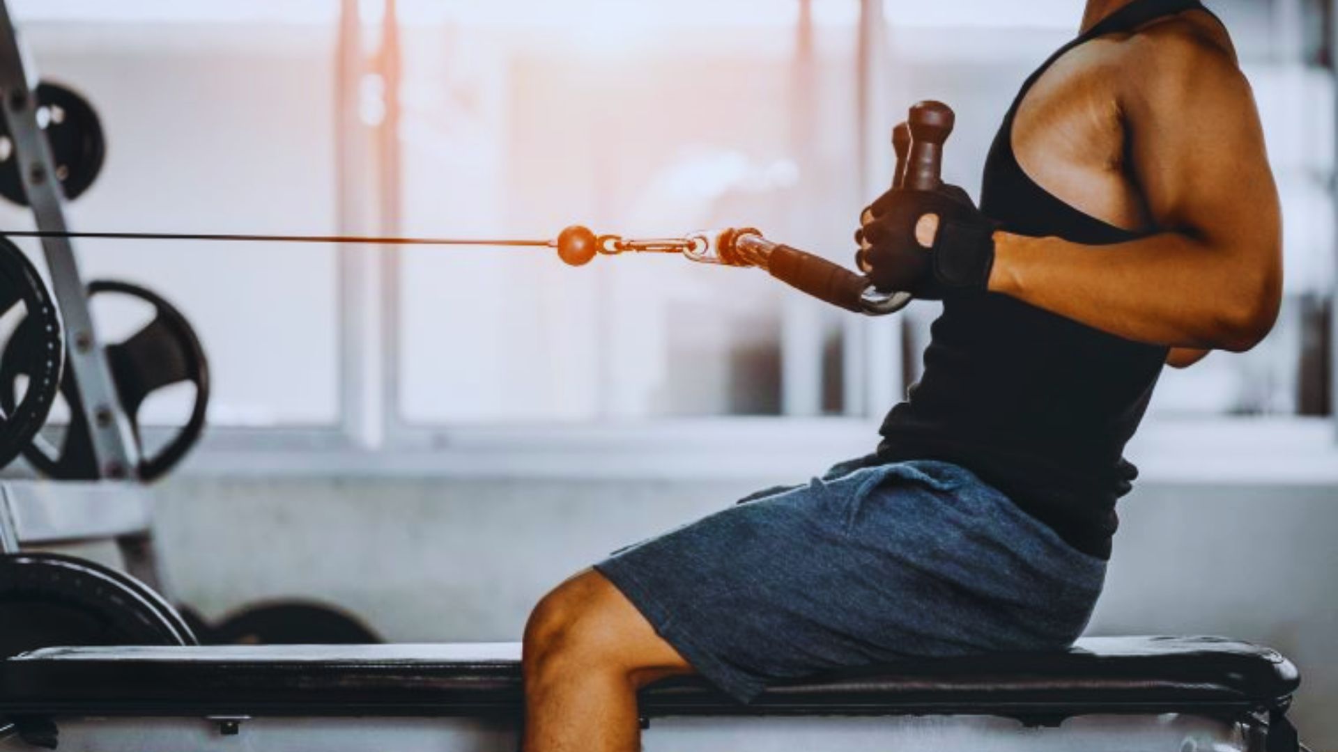 A person in athletic wear uses a seated cable row machine at the gym. Sunlight filters through windows, highlighting focused effort and strength.