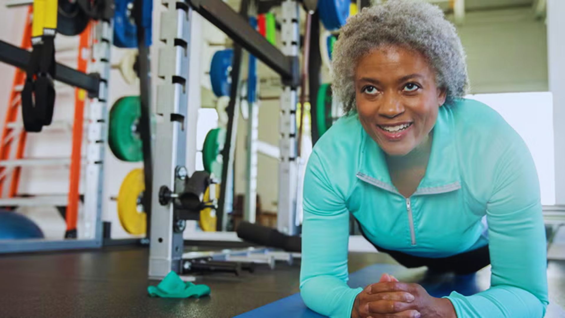 A woman with gray hair smiles confidently while doing a plank in a gym. She's on a blue mat, surrounded by weightlifting equipment. The atmosphere is energetic.