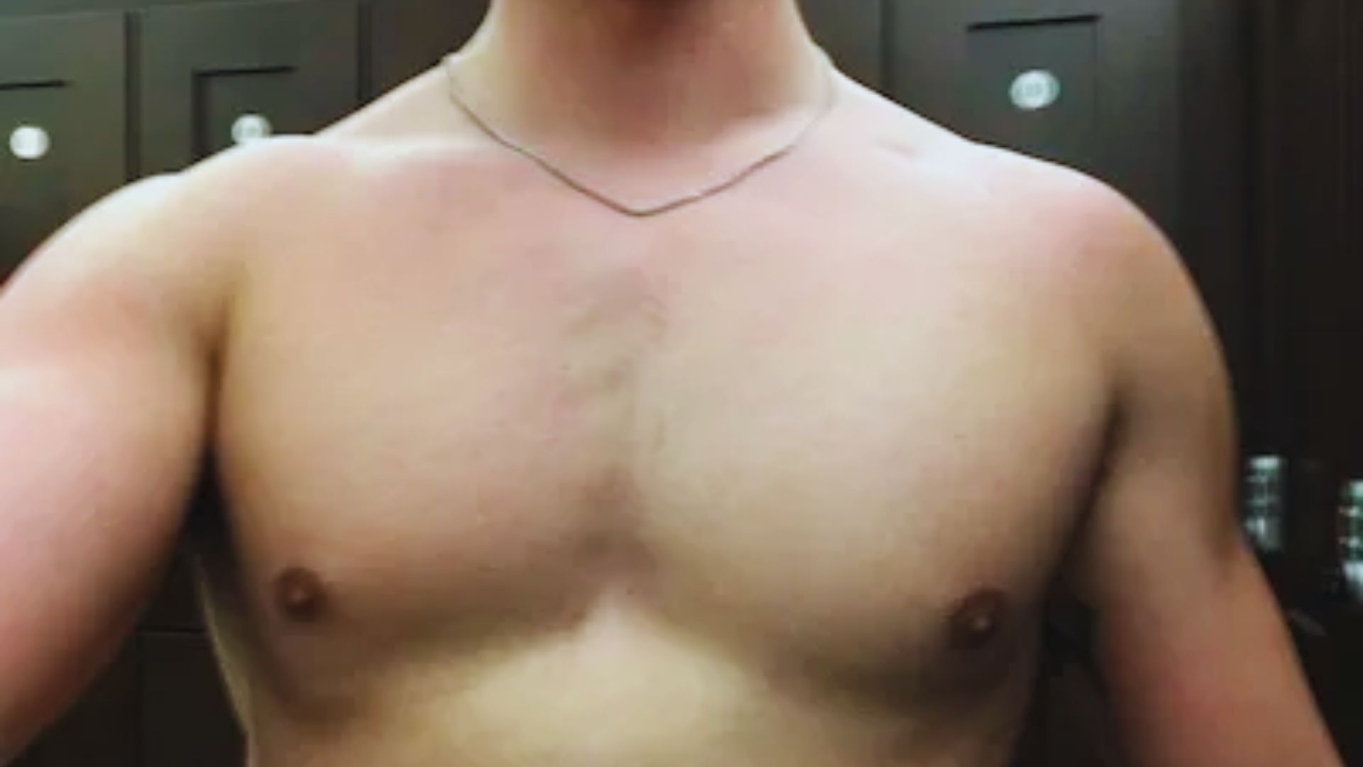 A shirtless person with a slight sunburn is seen in a gym locker room, wearing a thin chain necklace. Background features dark wooden lockers with round handle details.