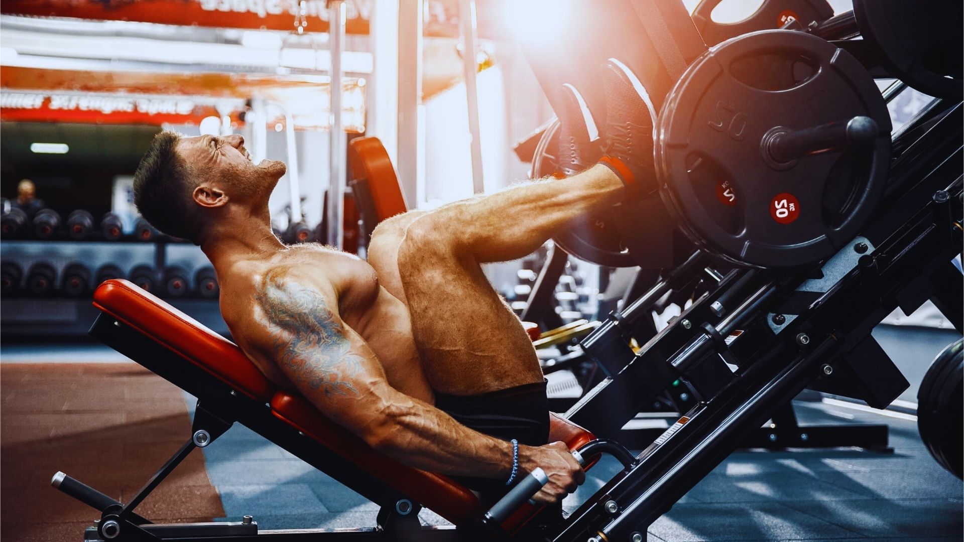 A muscular man intensely performs a leg press in a gym, showing strain and focus. The equipment is loaded with weights, emphasizing strength training.