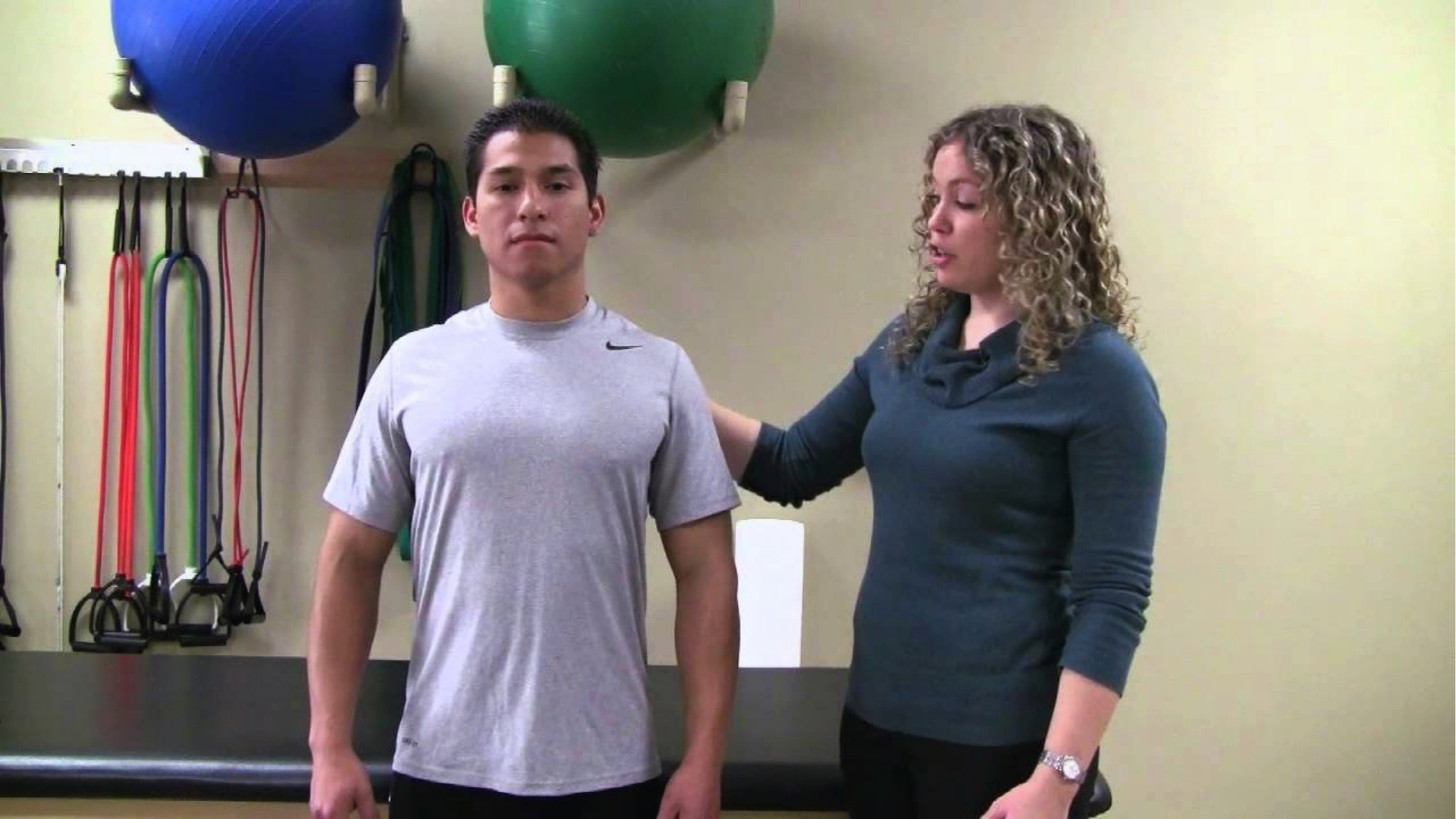 A woman with curly hair instructs a man in a grey shirt during a physiotherapy session. Exercise bands and therapy balls are visible in the background.