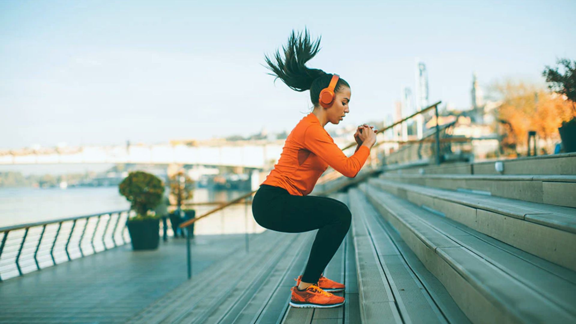A woman exercises on outdoor steps, wearing an orange outfit and headphones. Her hair is in motion, suggesting dynamic movement and focus.