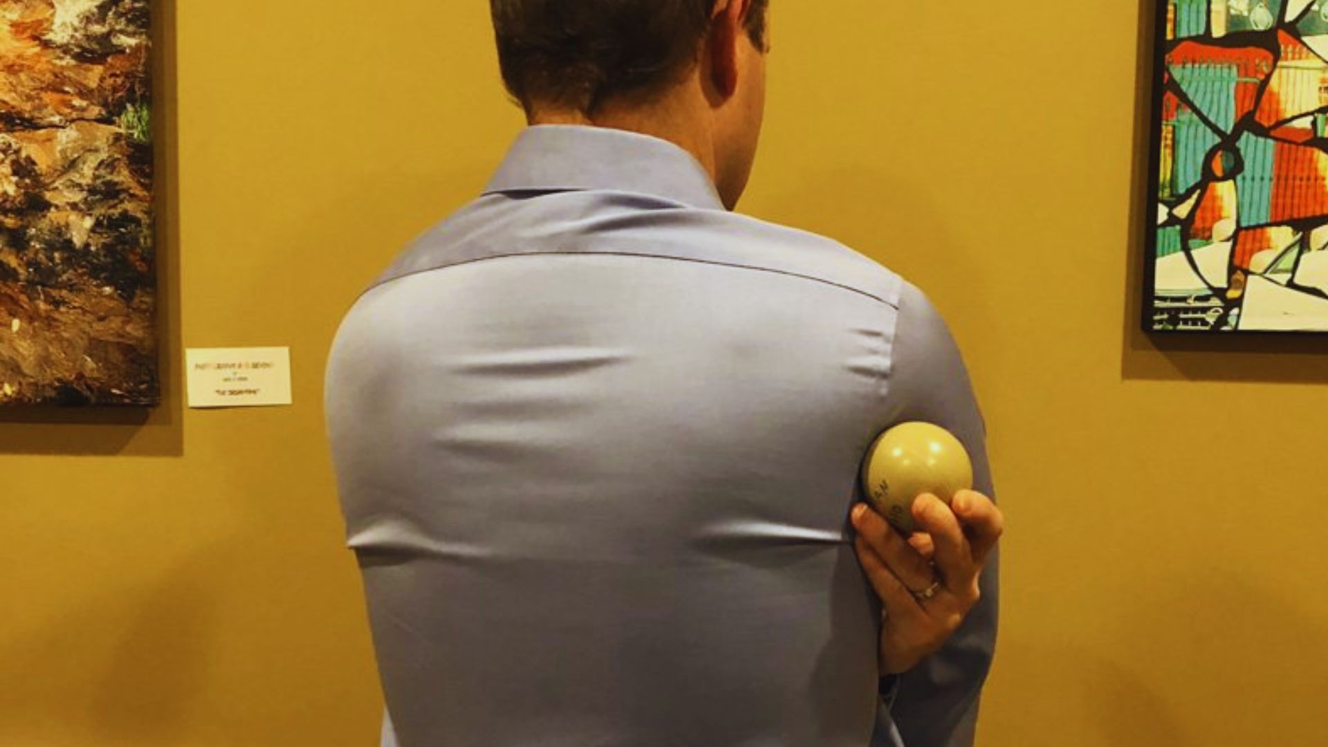 A person in a light blue shirt stands with their back to the camera, holding a billiard ball under their arm. Artworks are displayed on the wall around them.