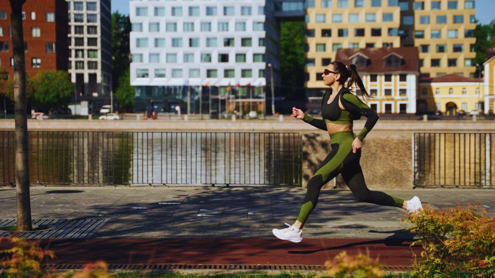 A woman in green athletic wear jogs energetically on a sunny day. She is on a riverside path with modern buildings in the background.