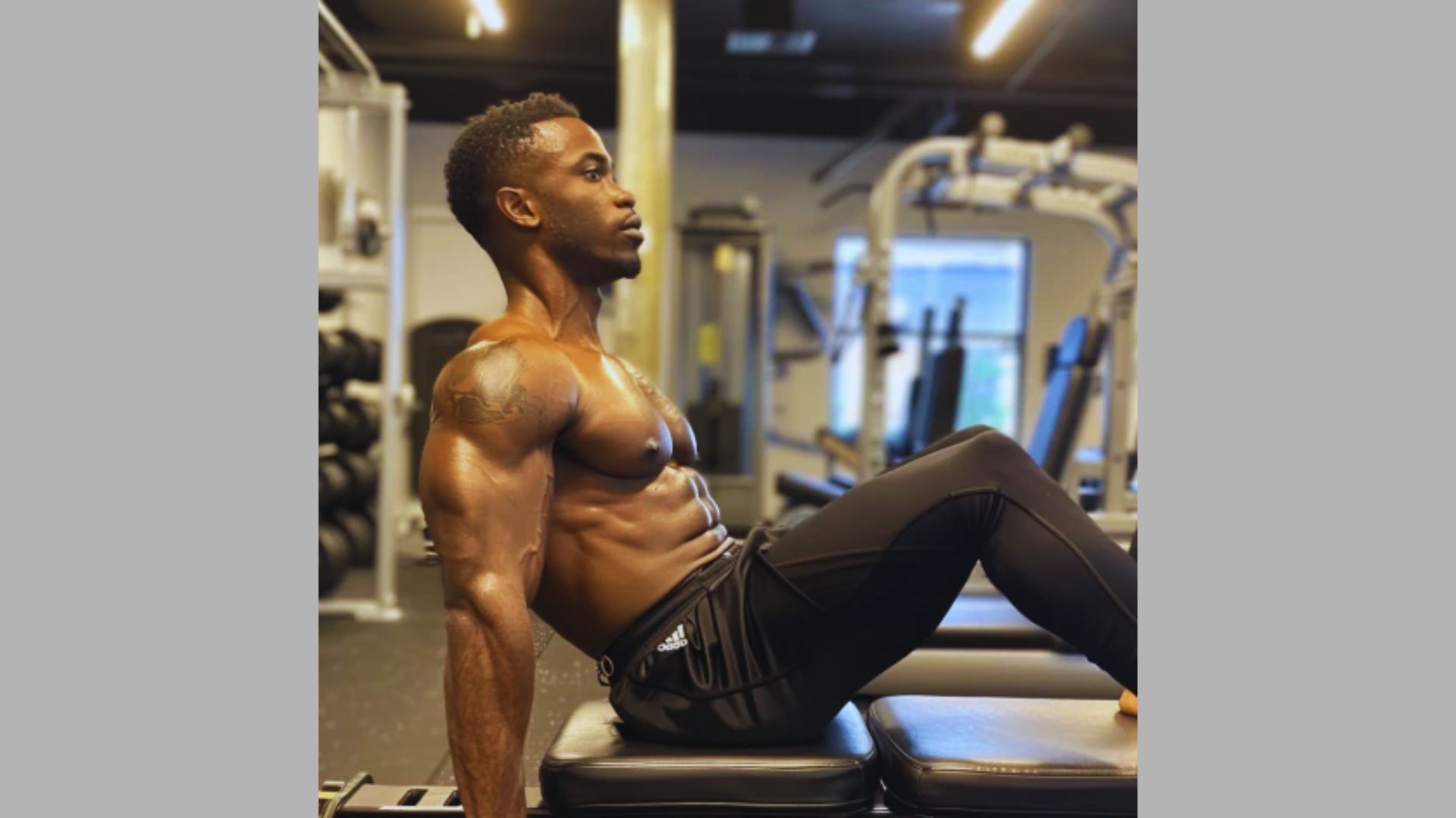 A fit person in a gym, sitting on a bench with arms supporting their weight behind them. They wear black athletic pants, showing a focused expression.