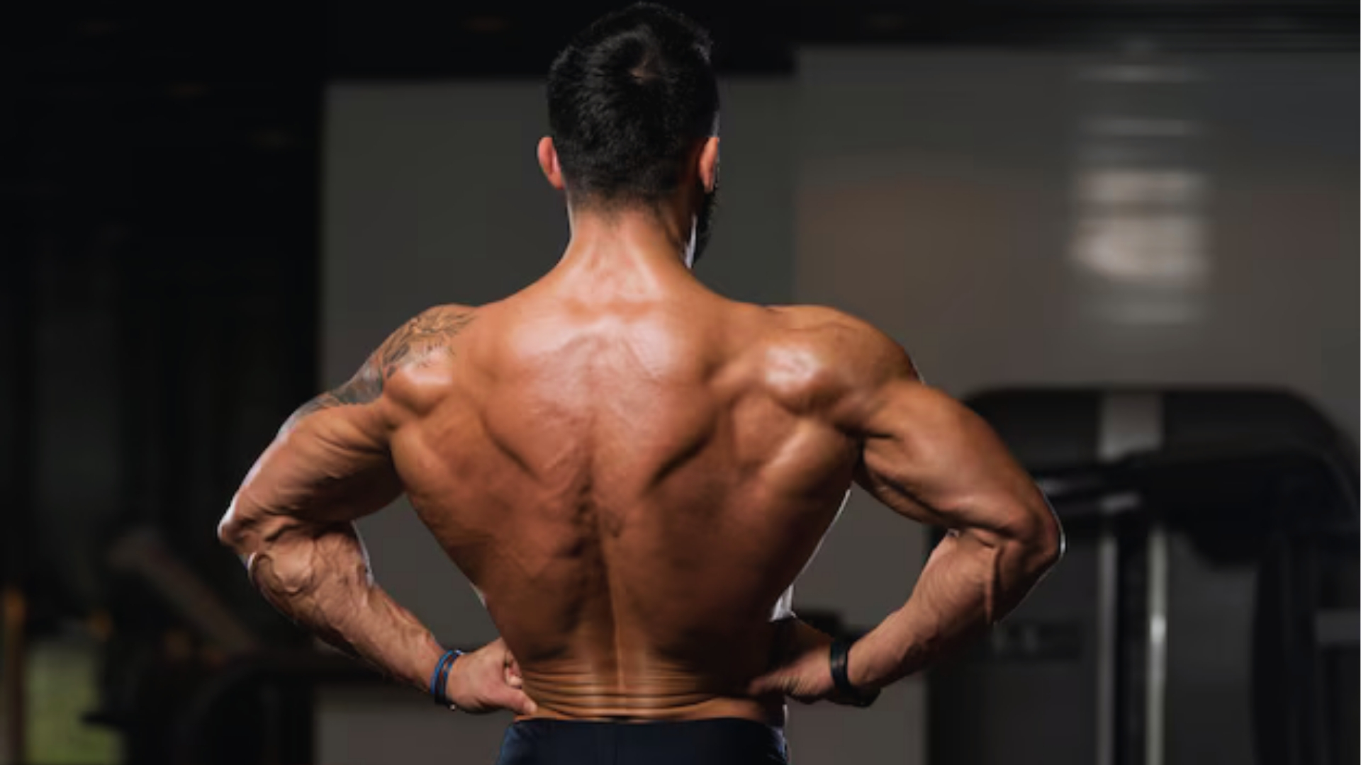 Muscular man viewed from the back in a gym setting, showcasing defined back and arm muscles. Dim lighting enhances the tone of strength and focus.