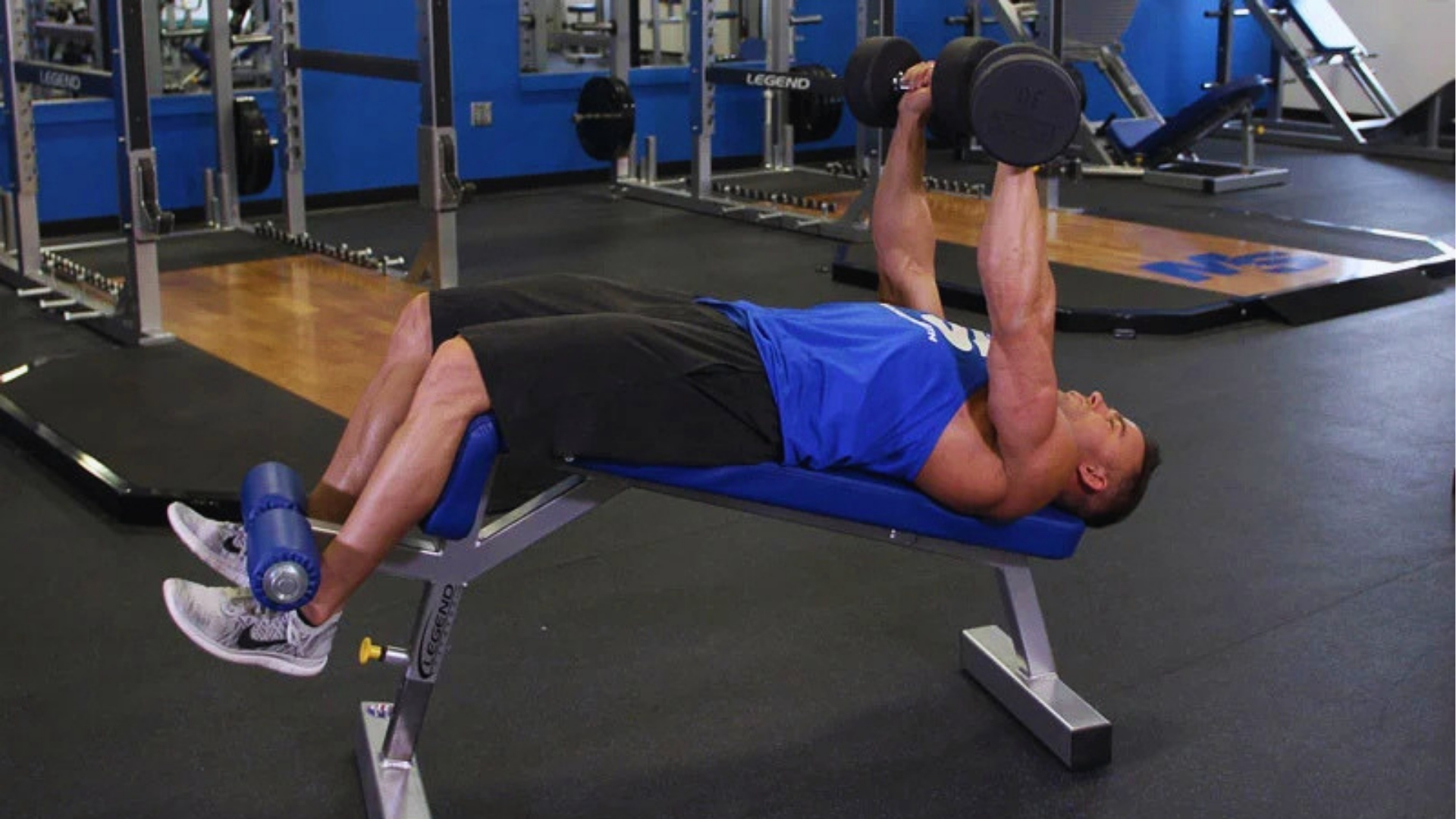 A person in athletic wear performs a dumbbell press on a blue bench in a gym. The space is equipped with weights and machines, creating a focused atmosphere.