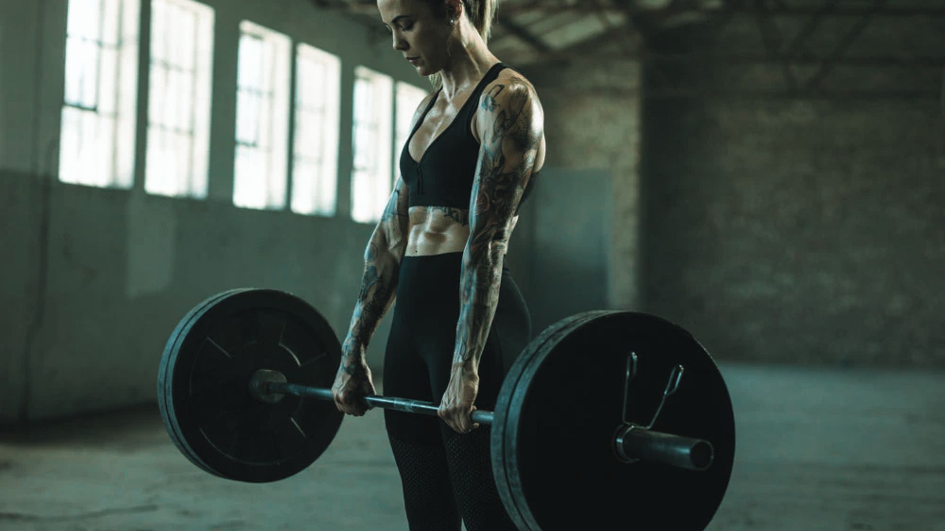A woman in athletic wear lifts a heavy barbell in an industrial-style gym. Sunlight streams through large windows, highlighting her toned muscles.