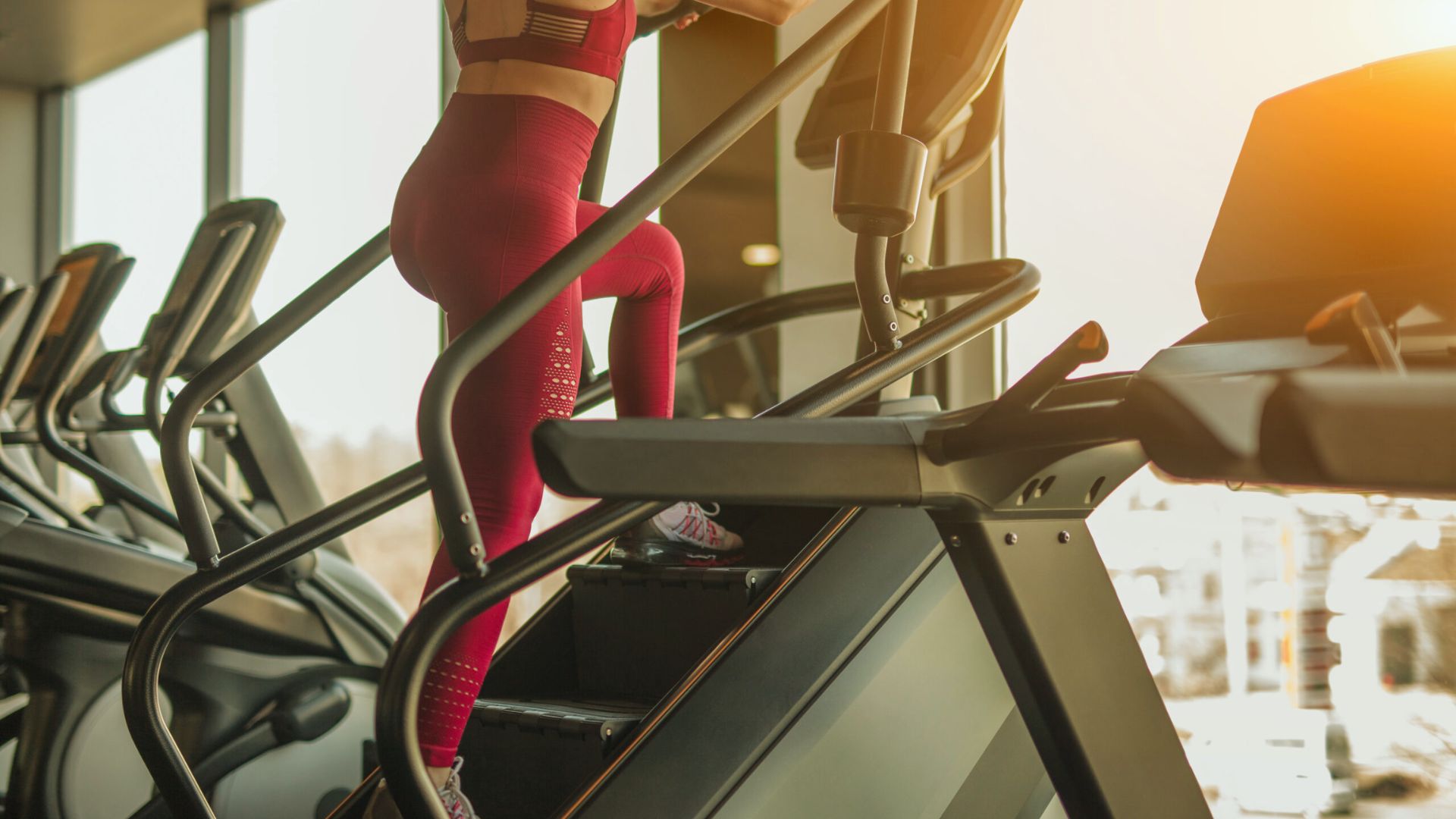 A person exercises on a stair climber in a gym, wearing red athletic wear. Sunlight streams through large windows, creating a warm, energetic atmosphere.