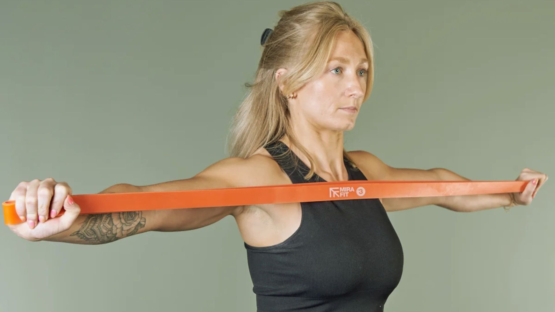 A woman in workout attire stretches an orange resistance band across her chest. Her expression is focused, conveying determination and strength.