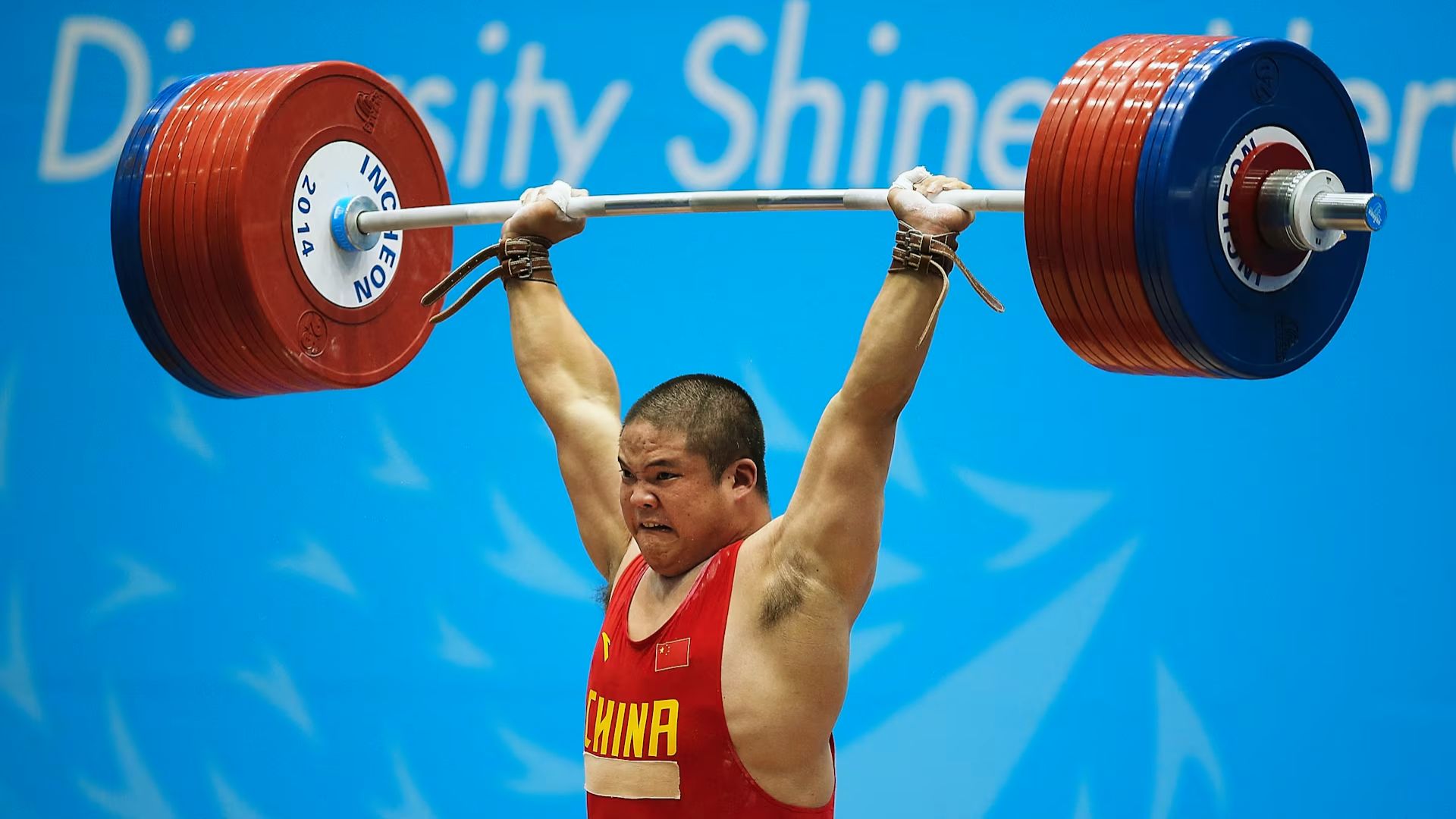 A weightlifter in a red uniform labeled "China" lifts a heavy barbell overhead, showing intense focus and strength. The background is a bright blue with white text.