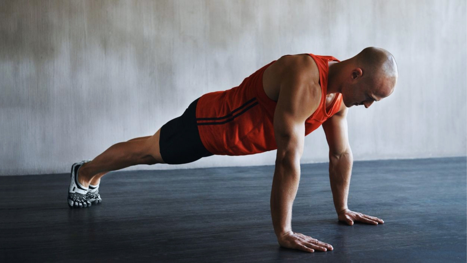 A person in a red tank top and black shorts performing a plank on a dark floor. The setting is minimal, conveying focus and determination.