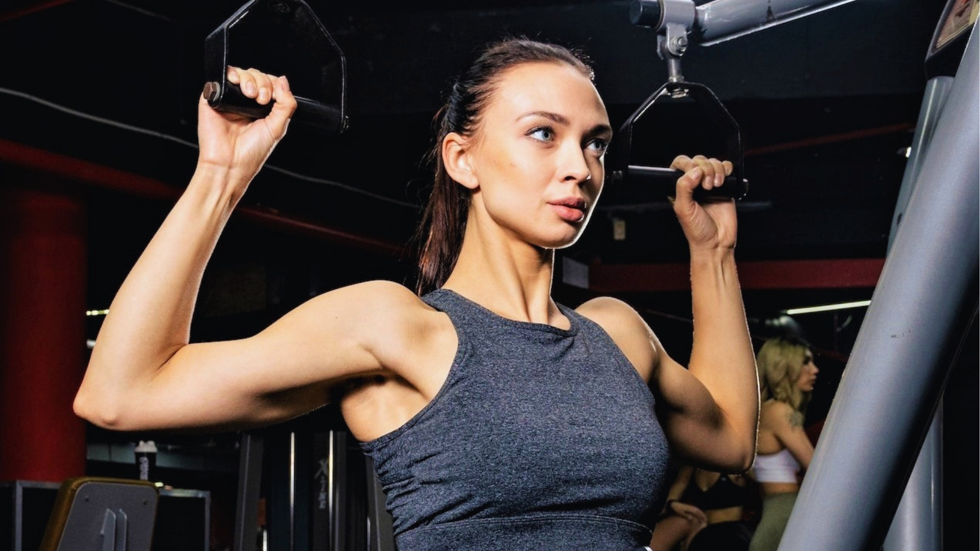 A woman in a gym uses a lat pulldown machine, wearing a gray tank top. Her expression is focused and determined, capturing a sense of strength.