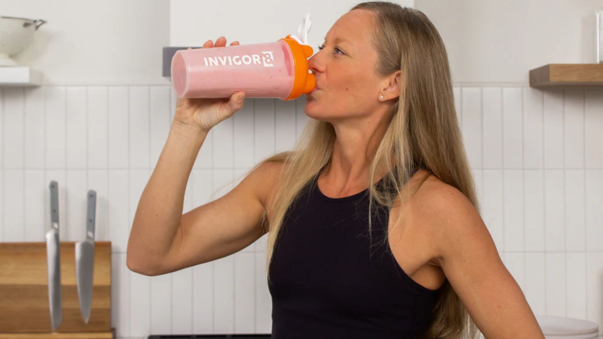 A woman sipping a smoothie directly from a blender in a modern kitchen setting.