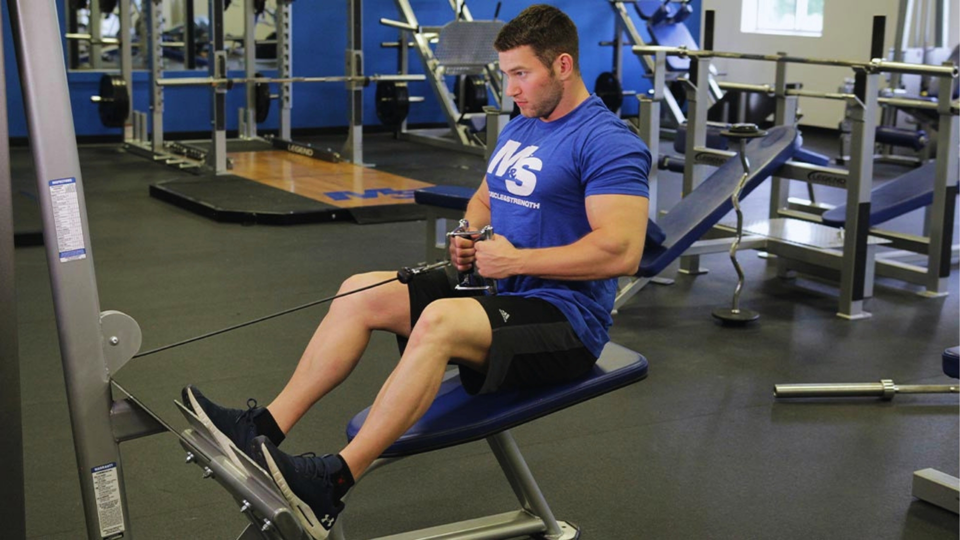 A person in a blue T-shirt and black shorts is focused on using a seated cable row machine in a gym. The setting is organized and well-equipped.