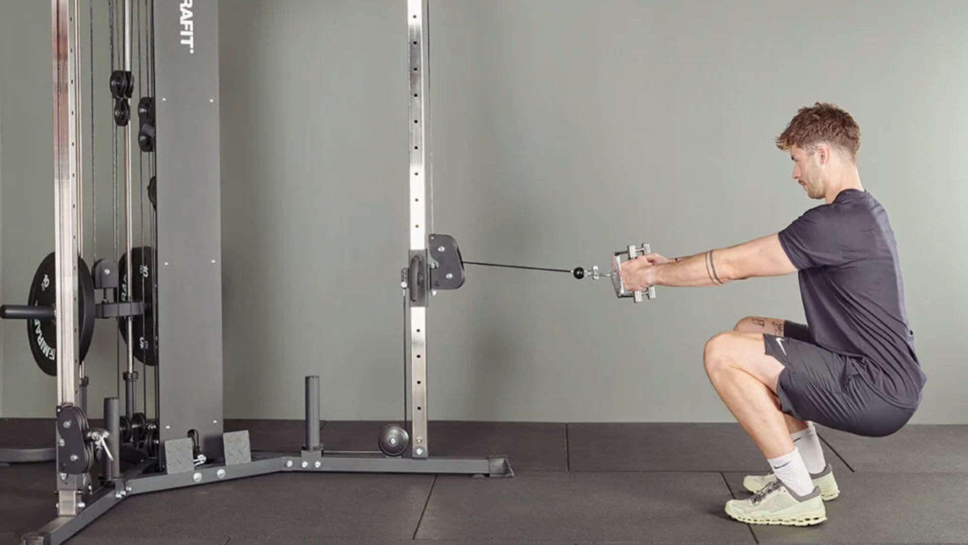 A man in athletic wear performs a low cable squat, holding a cable machine handle. He is focused, demonstrating strength and balance on a gym mat.