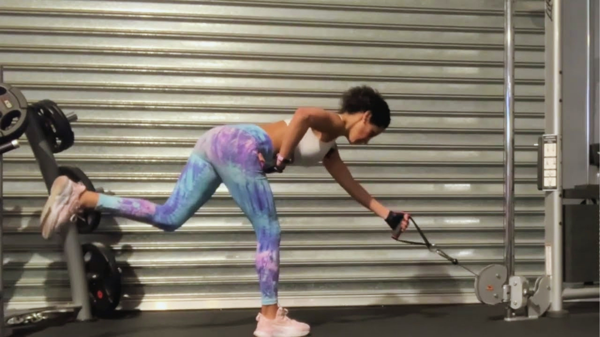A woman exercises in a gym using a cable machine. She's wearing colorful leggings and a white top, focusing intensely during her workout.