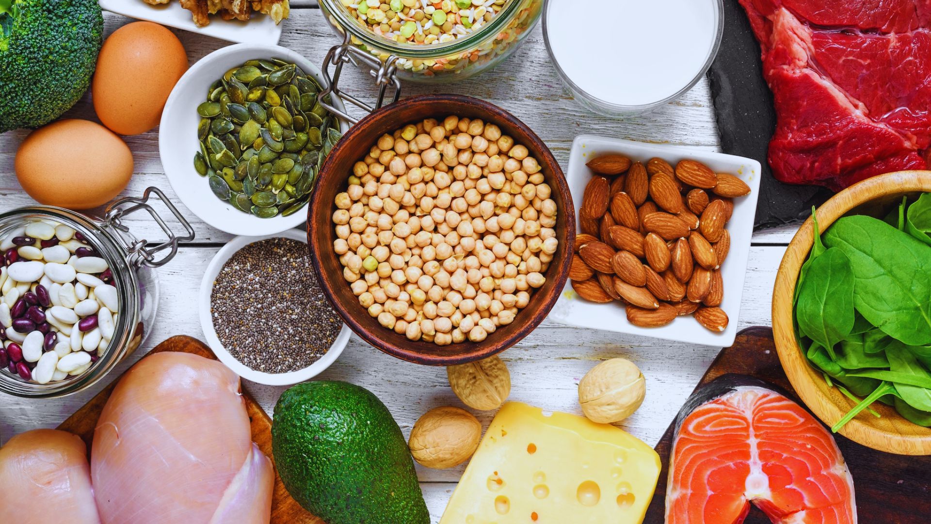 A vibrant assortment of foods on a white wooden table, including chickpeas, almonds, spinach, salmon, cheese, avocado, chicken, eggs, seeds, and beans.