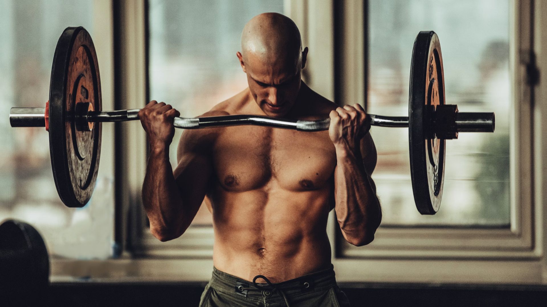 A muscular, bald man intensely lifts a barbell indoors. He is shirtless, highlighting his defined physique. The atmosphere is focused and determined.