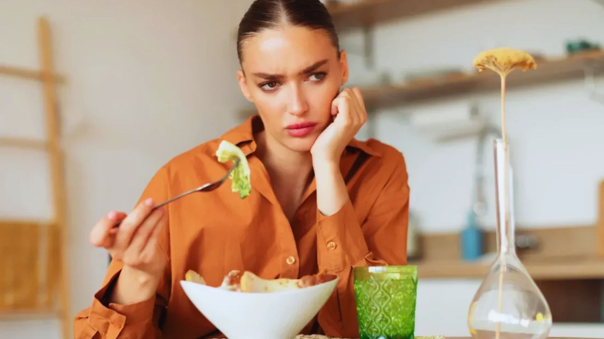 A woman in an orange shirt looks displeased while holding a fork with a typical salad over a bowl. A green glass and vase are on the table. The tone is contemplative.