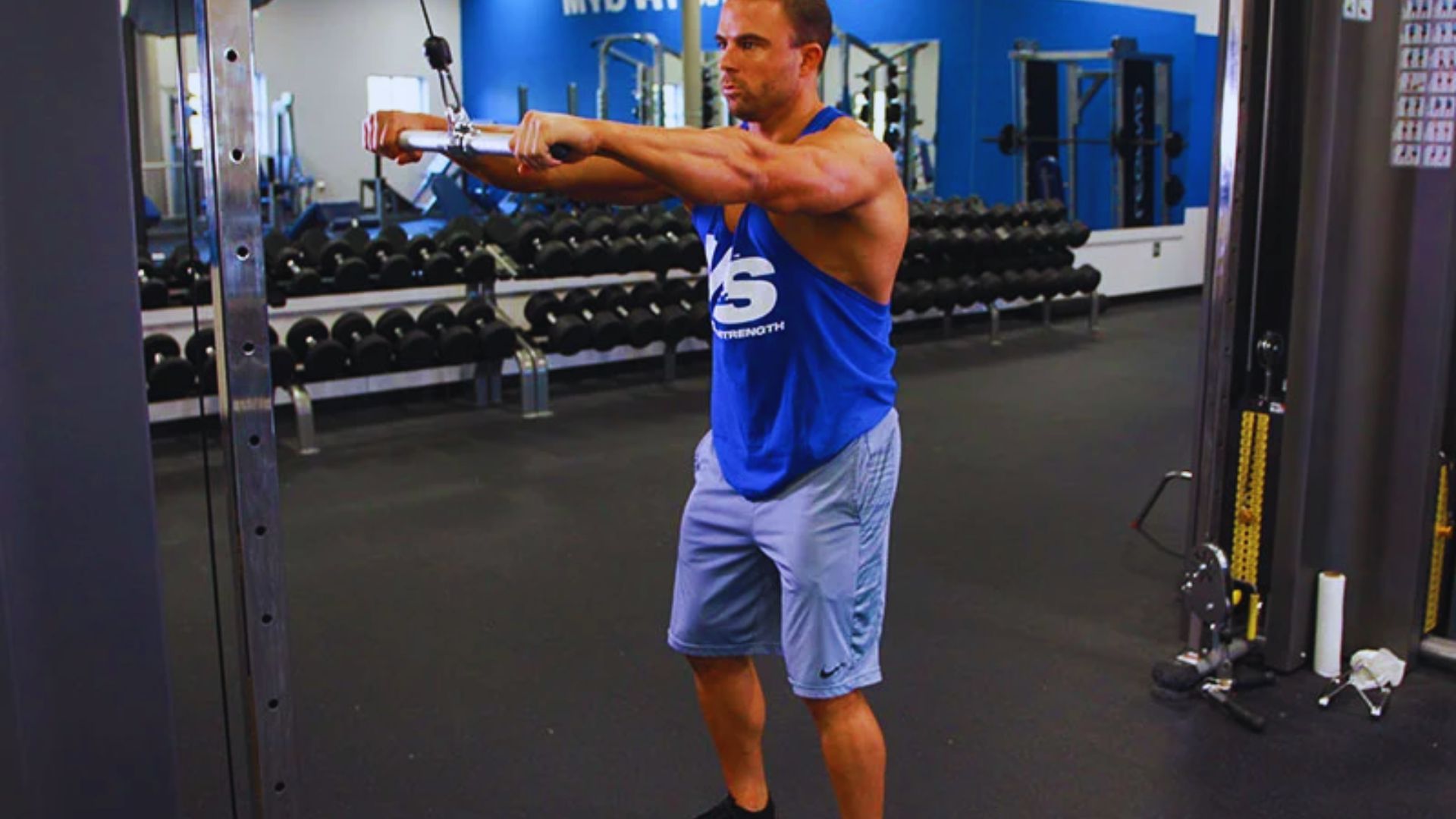 Man in a blue tank top exercises with a cable machine in a gym. He stands focused, arms extended forward, surrounded by weights in a brightly lit space.