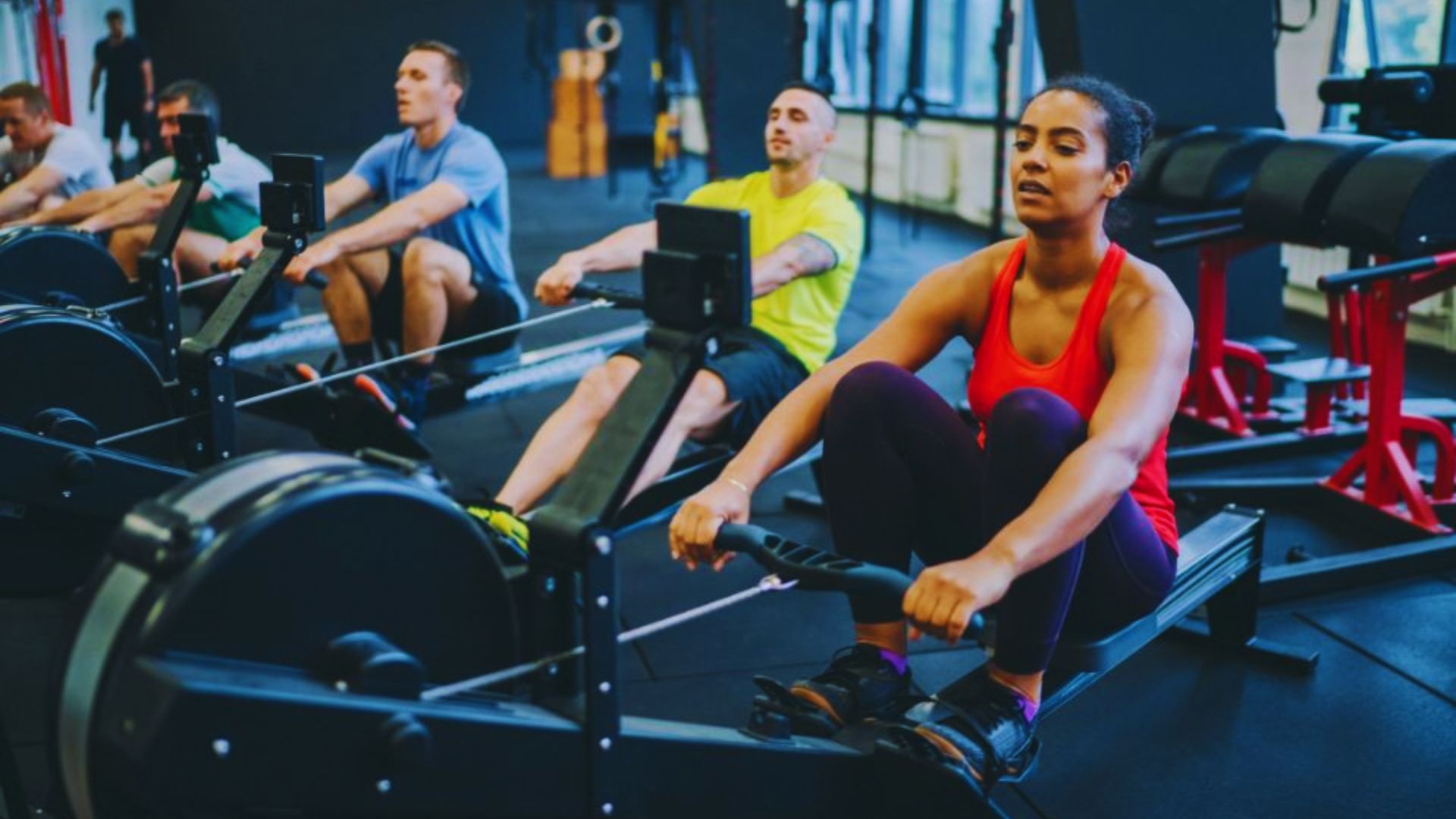 A focused group of people exercises on rowing machines in a gym. The front woman wears a red tank top, creating a dynamic and energetic atmosphere.