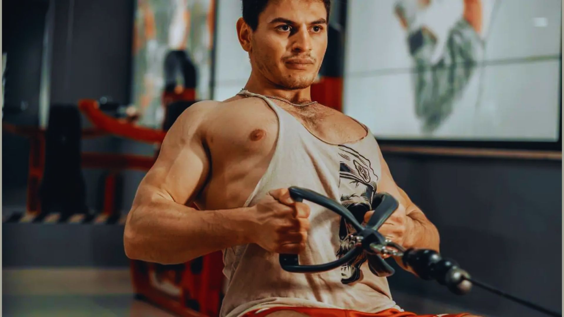 A man intensely exercises on a rowing machine in a gym, wearing a sleeveless shirt. The focus is on his muscular build and determined expression.