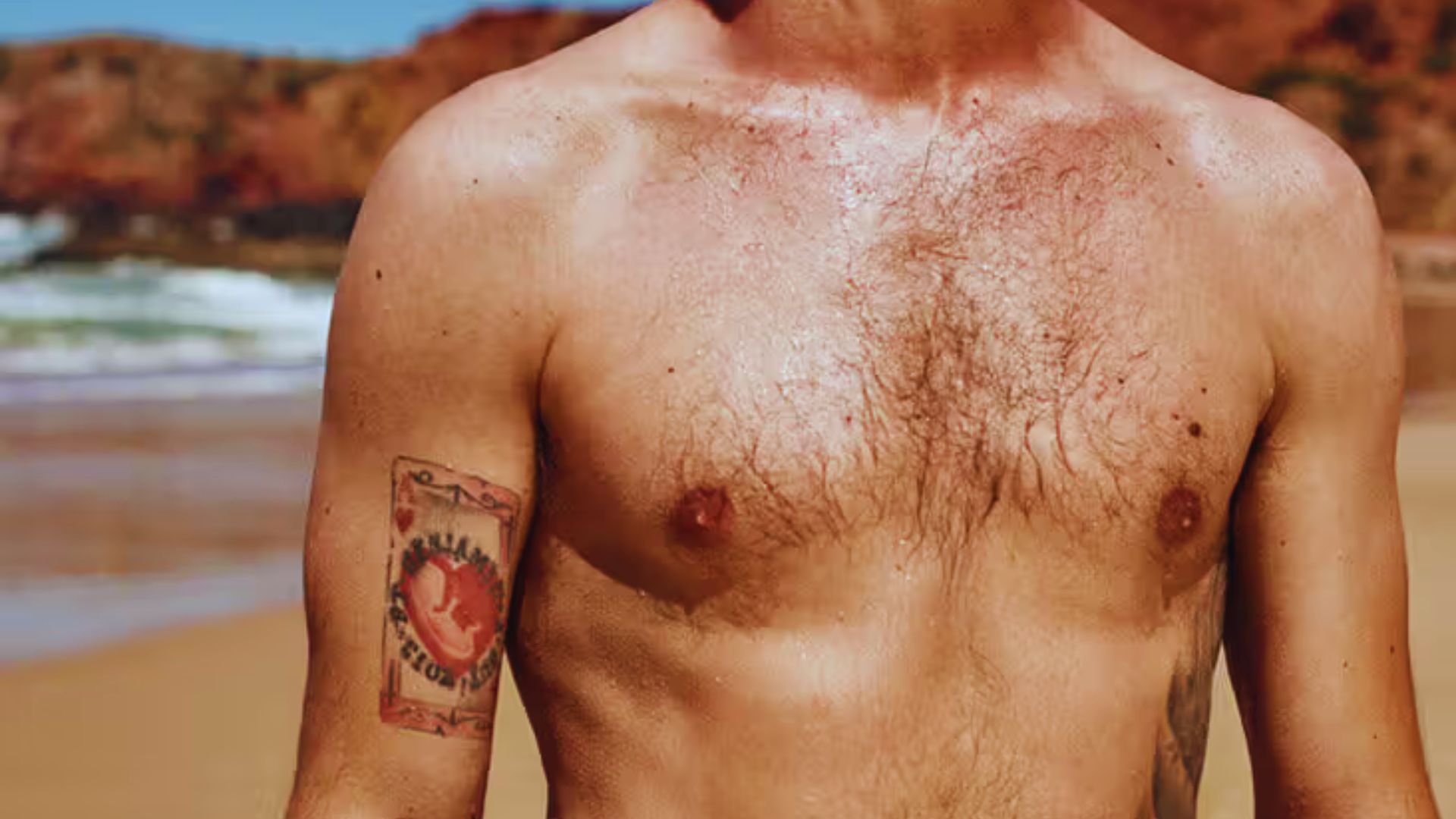 A shirtless man with chest hair and a tattoo on his arm stands on a sunny beach. The background features sand, ocean waves, and rocky cliffs.