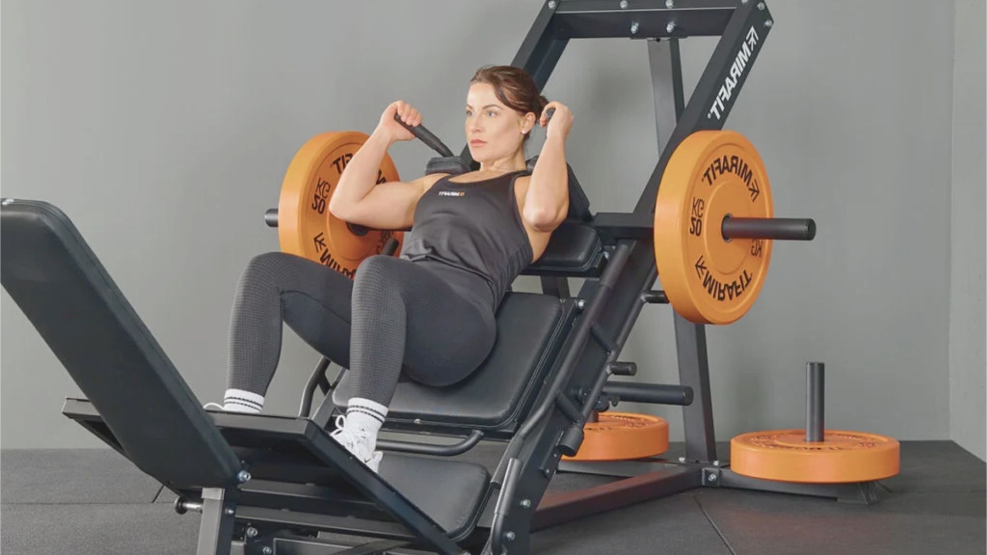 A woman in workout attire uses a leg press machine with orange weights in a gym. She looks focused and determined, conveying strength and motivation.
