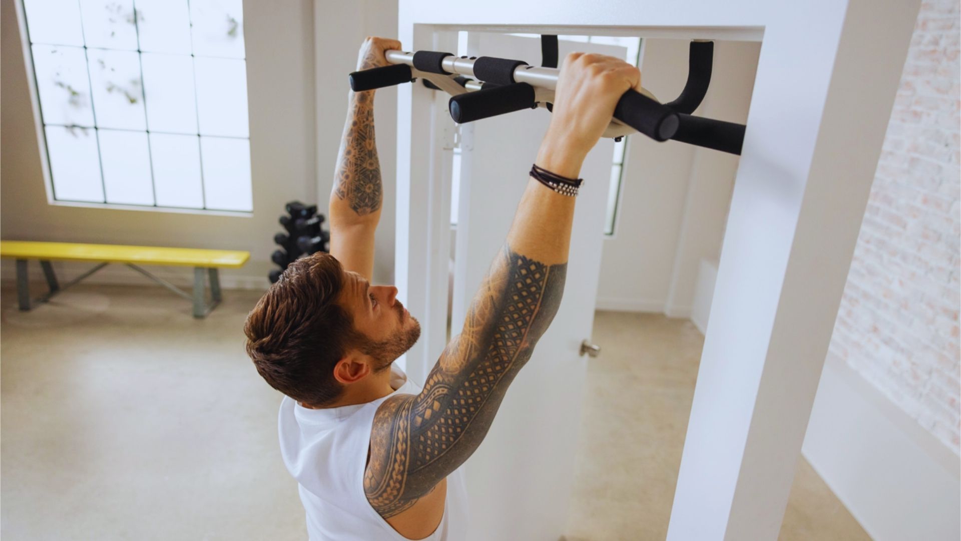 A man with tattooed arms does pull-ups on a doorway pull-up bar in a bright room, conveying focus and strength. A window and weights are in the background.