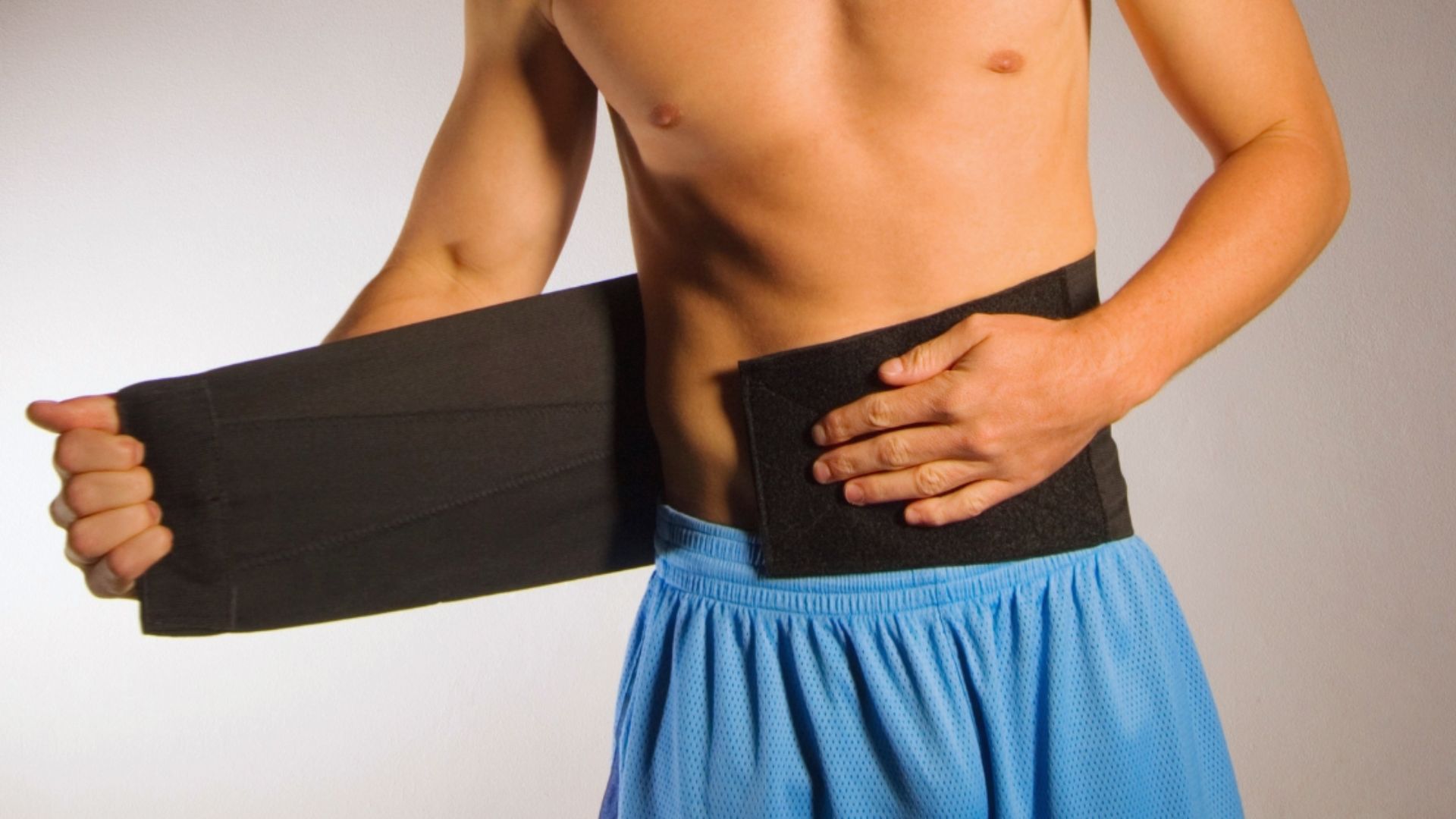 A shirtless person wearing blue shorts adjusts a black abdominal belt around their waist. The setting is bright, conveying a sense of fitness and wellbeing.