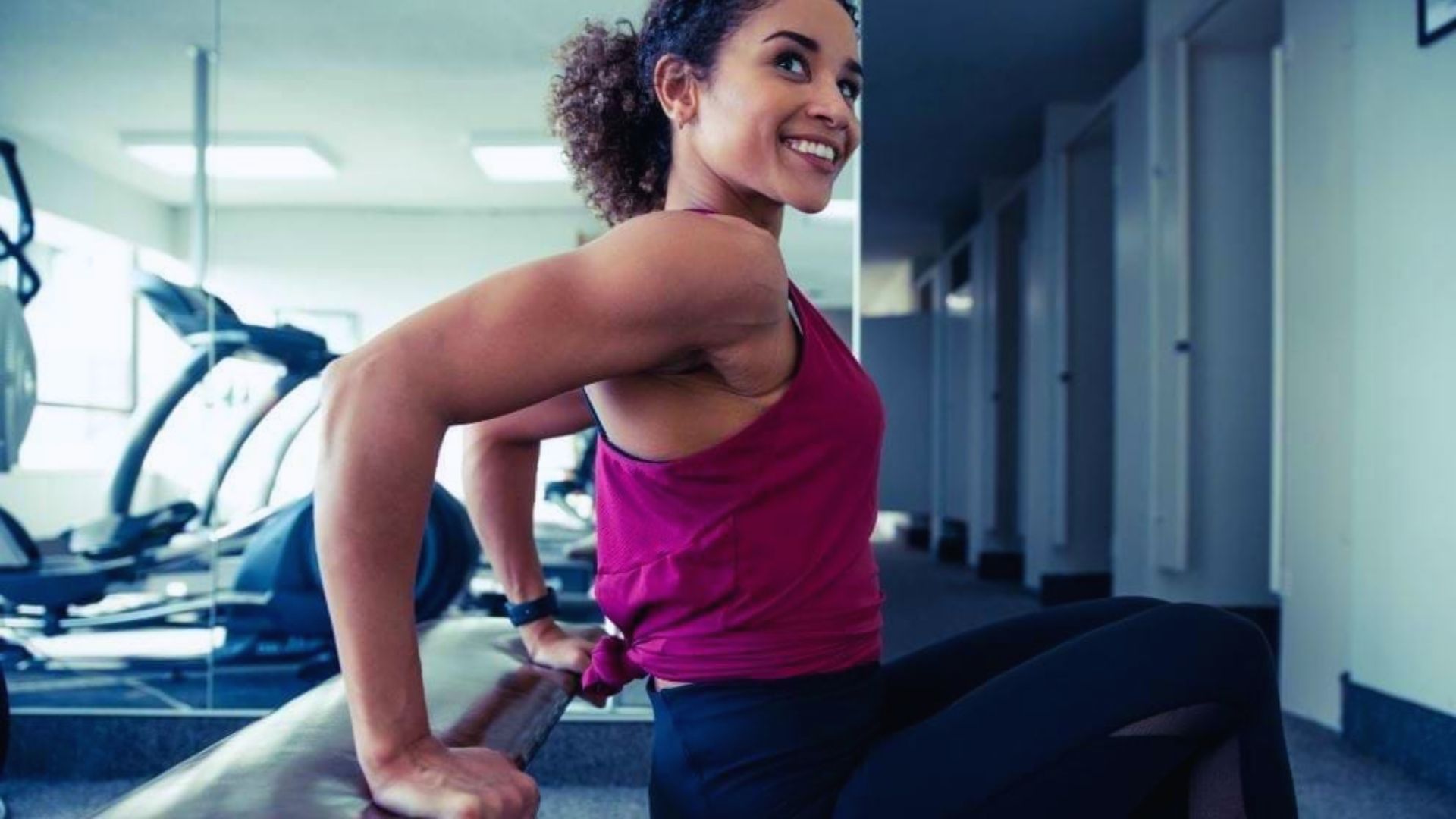 Woman in a pink tank top smiling while doing a tricep dip on a bench in a gym. The room is bright, with exercise machines in the background.
