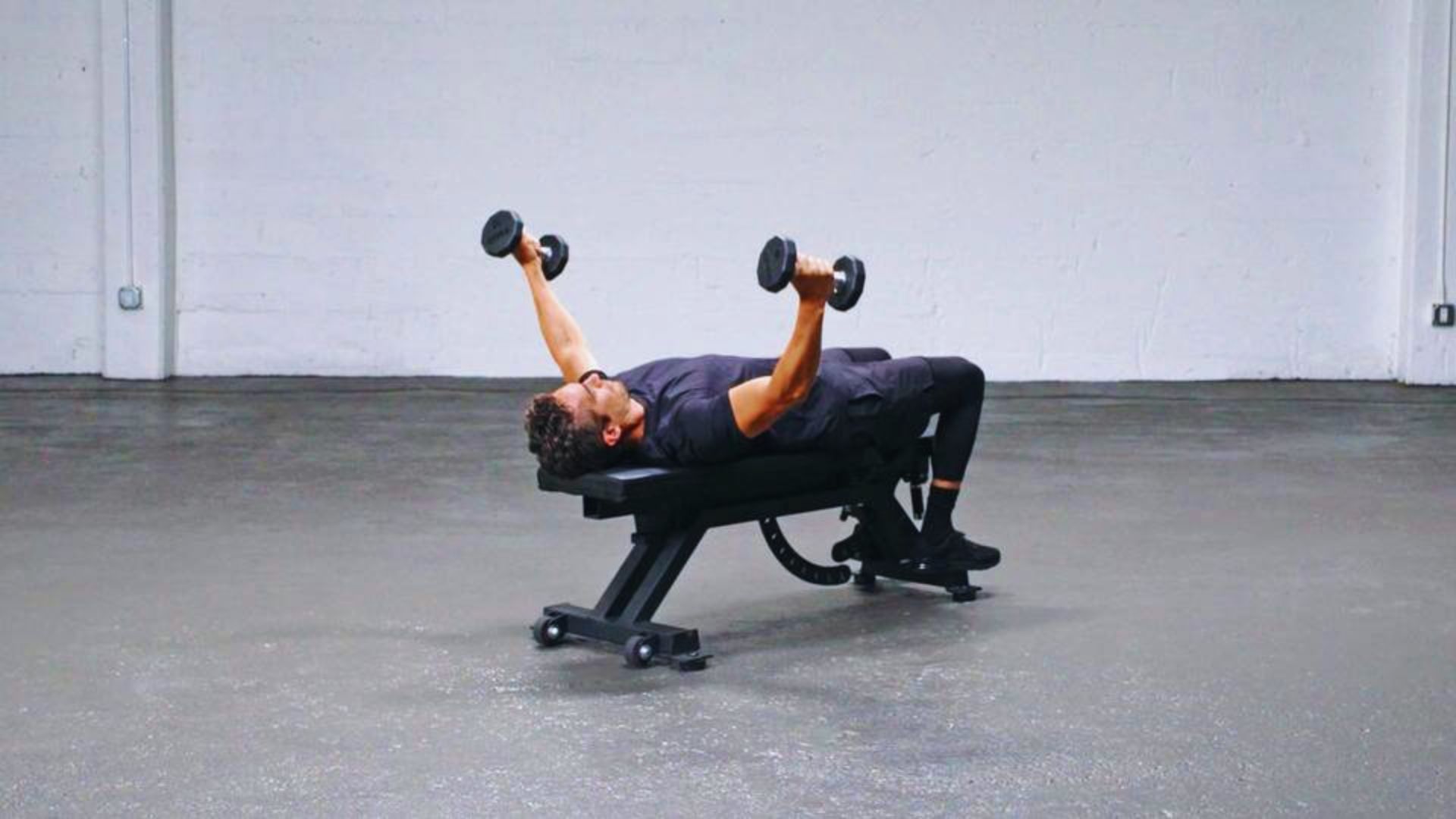 A person is performing a chest press on a bench with dumbbells in each hand. They are in a spacious, minimalist gym with a focused expression.