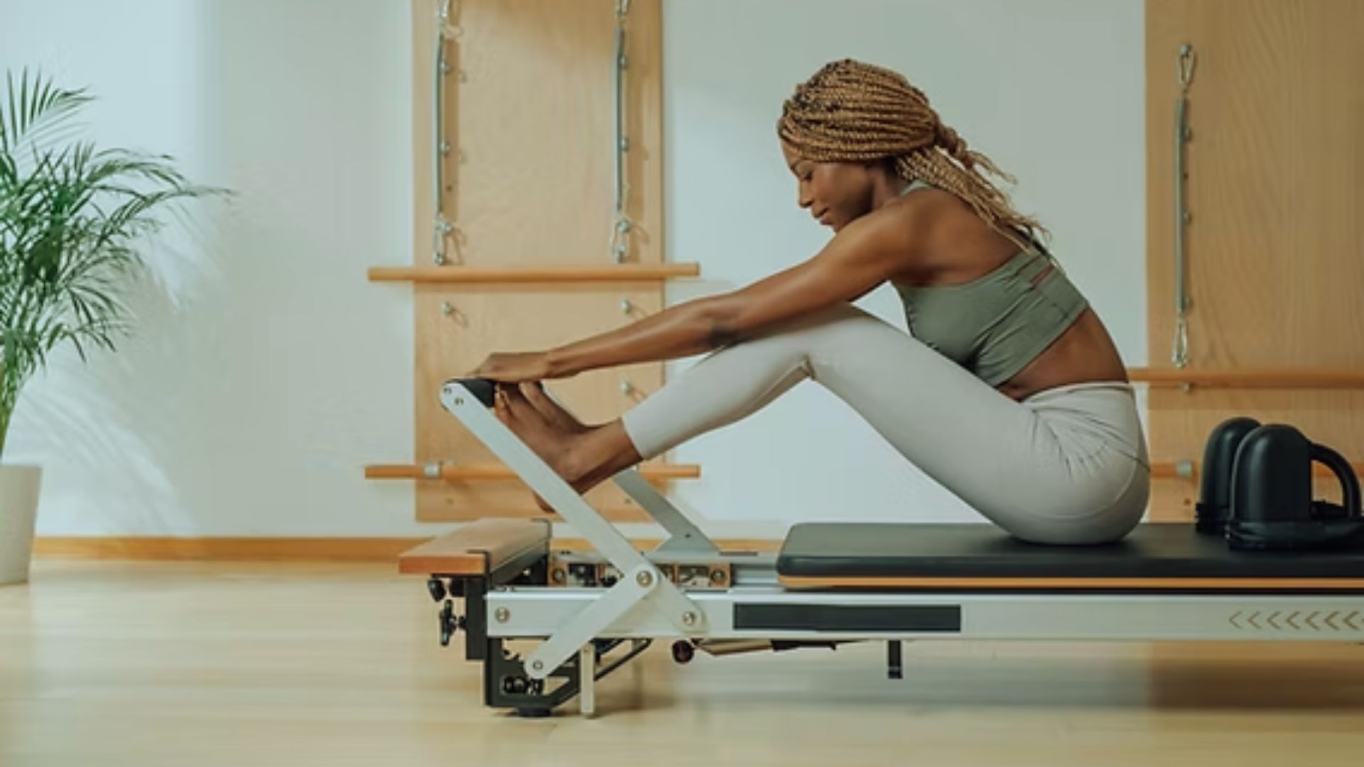 A woman with braided hair does a Pilates stretch on a reformer in a bright room. She wears a green sports bra and gray leggings, creating a calm, focused atmosphere.