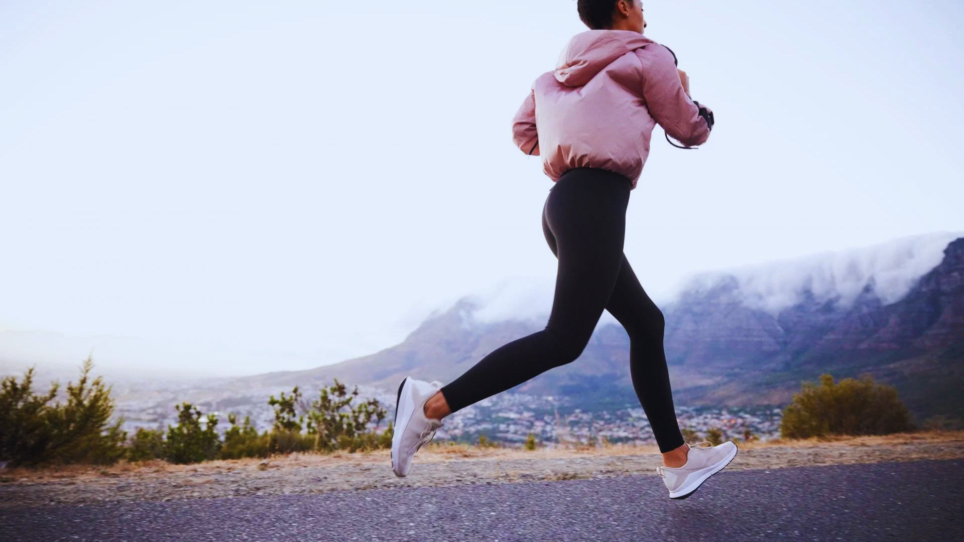 A person in athletic wear jogs on a road, wearing a pink jacket and black leggings. Mountains with mist rise in the background under a clear sky.