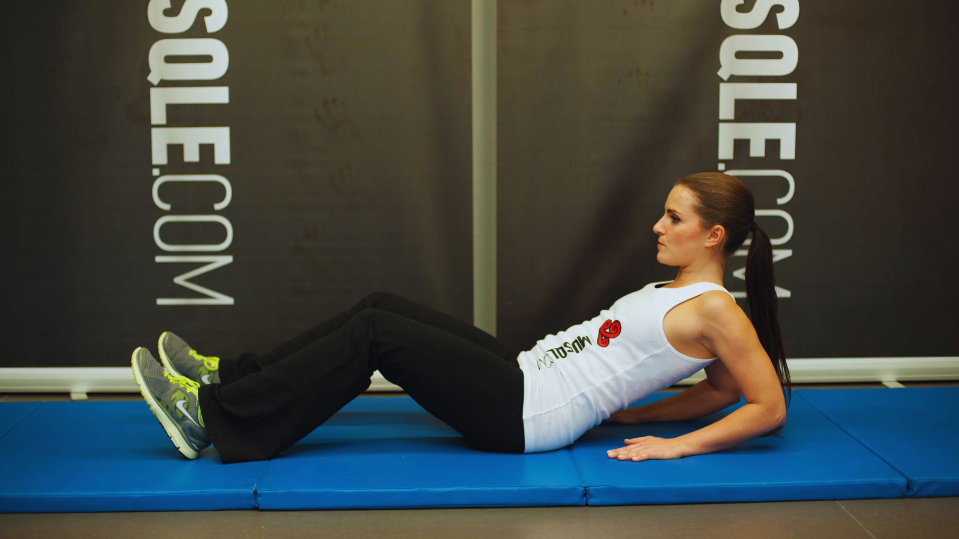 A woman in workout attire performs an abdominal exercise on a blue mat in a gym. She is lying on her back with her knees bent, conveying focus.