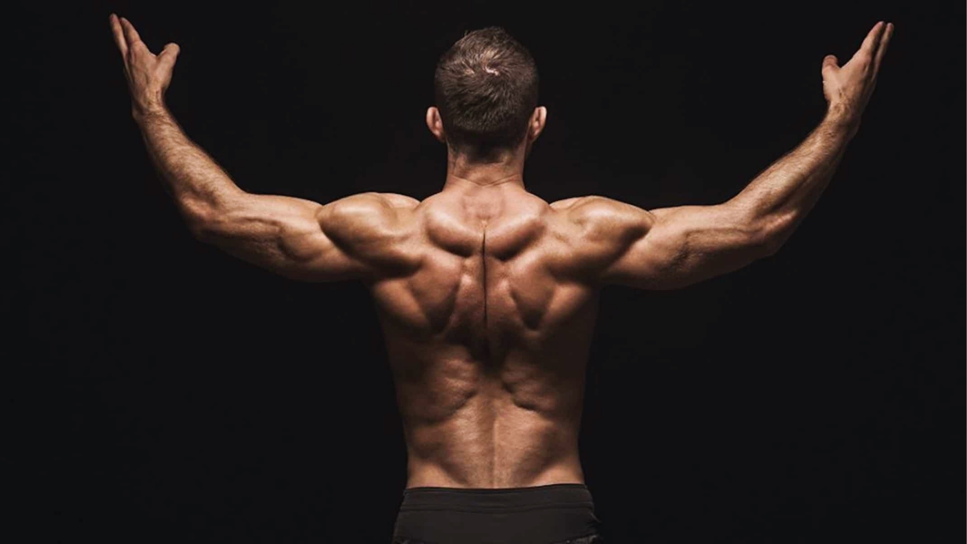 Muscular man with arms raised, back view. The lighting highlights his defined muscles against a dark background, conveying strength and power.