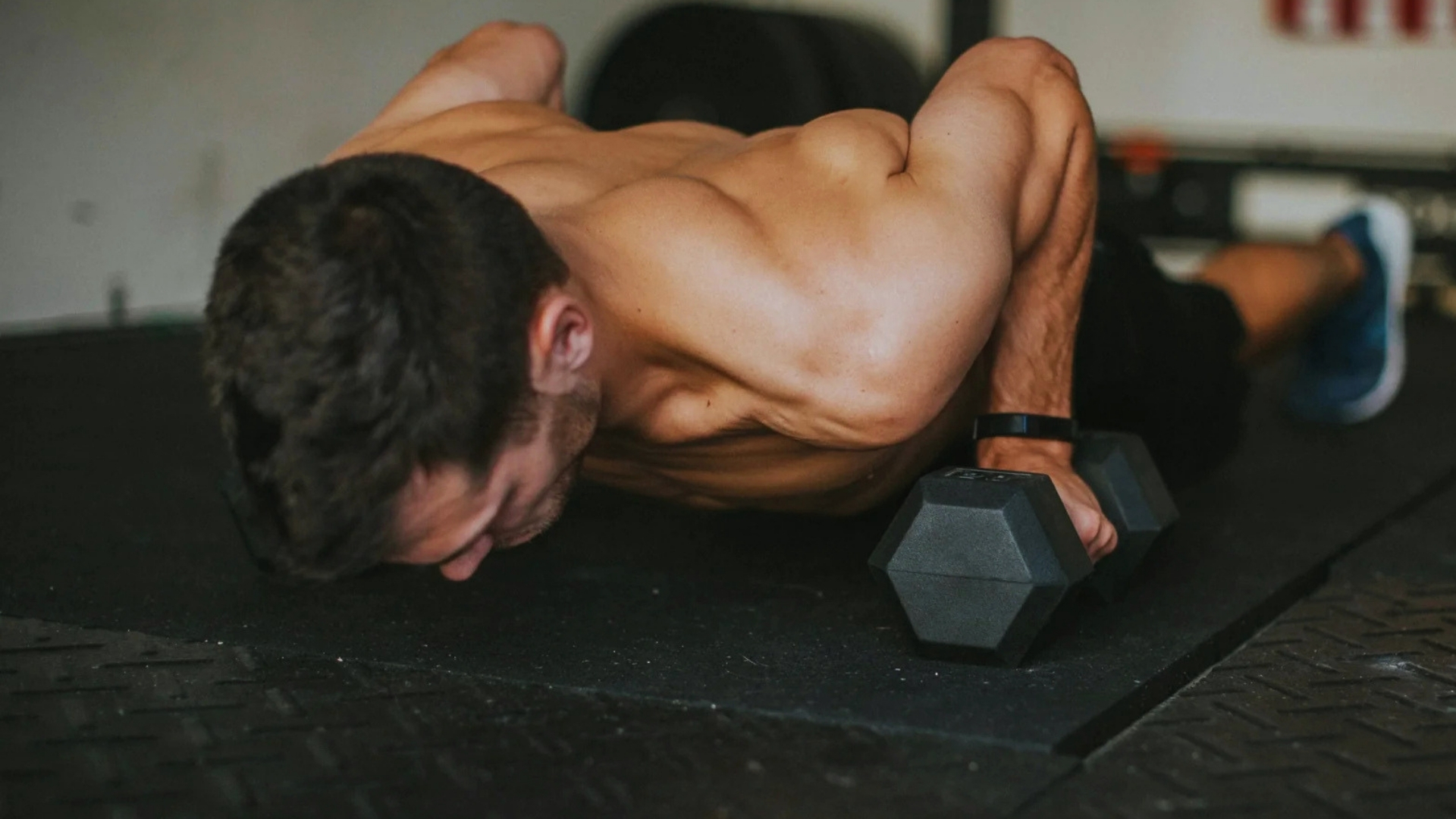 Man in a gym performing push-ups with dumbbells, showcasing muscular torso. The setting conveys strength and focus.