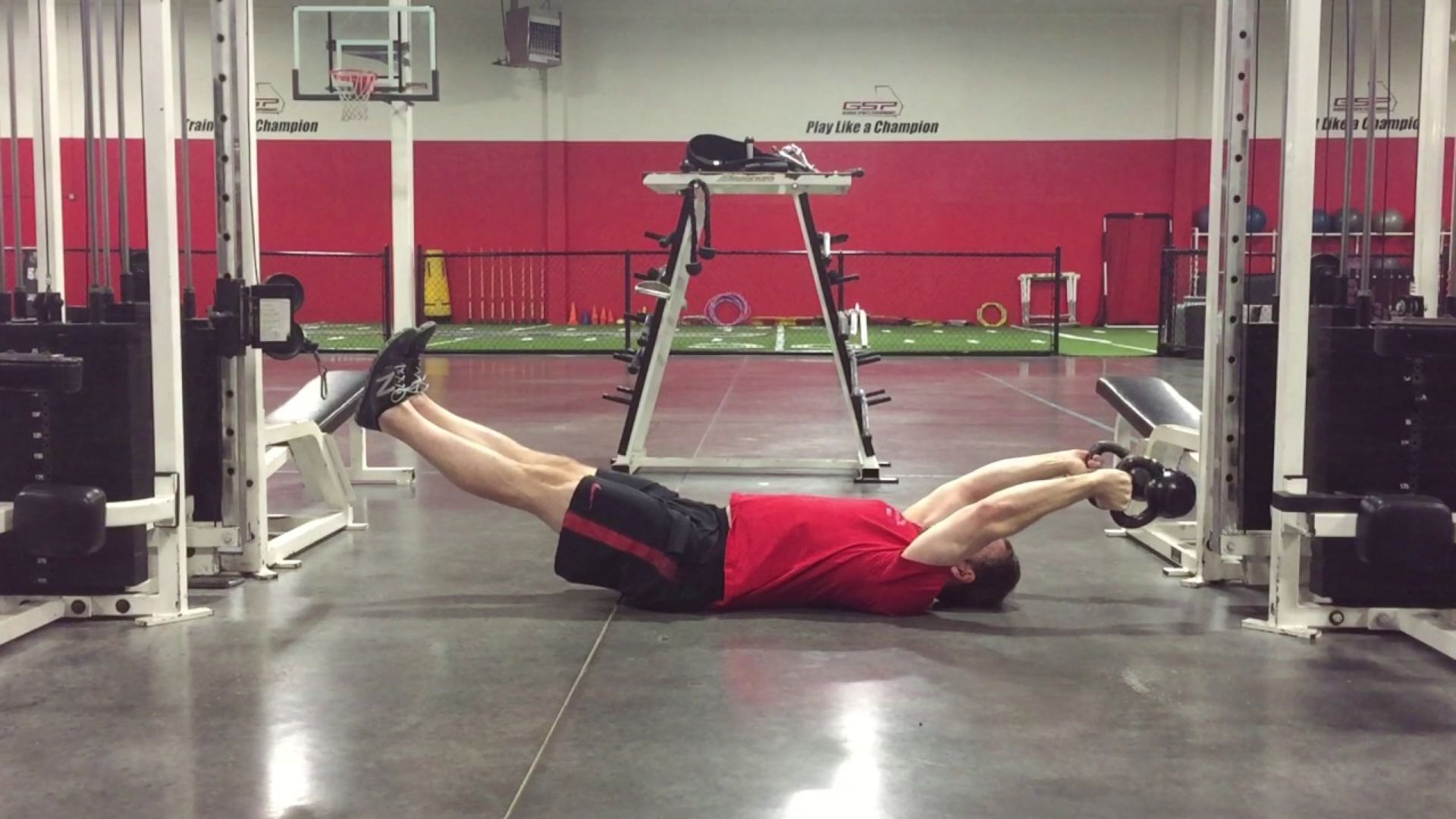 A person in a red shirt exercises in a gym, lying on their back holding a weight with outstretched arms. The gym has red and white walls and various equipment.