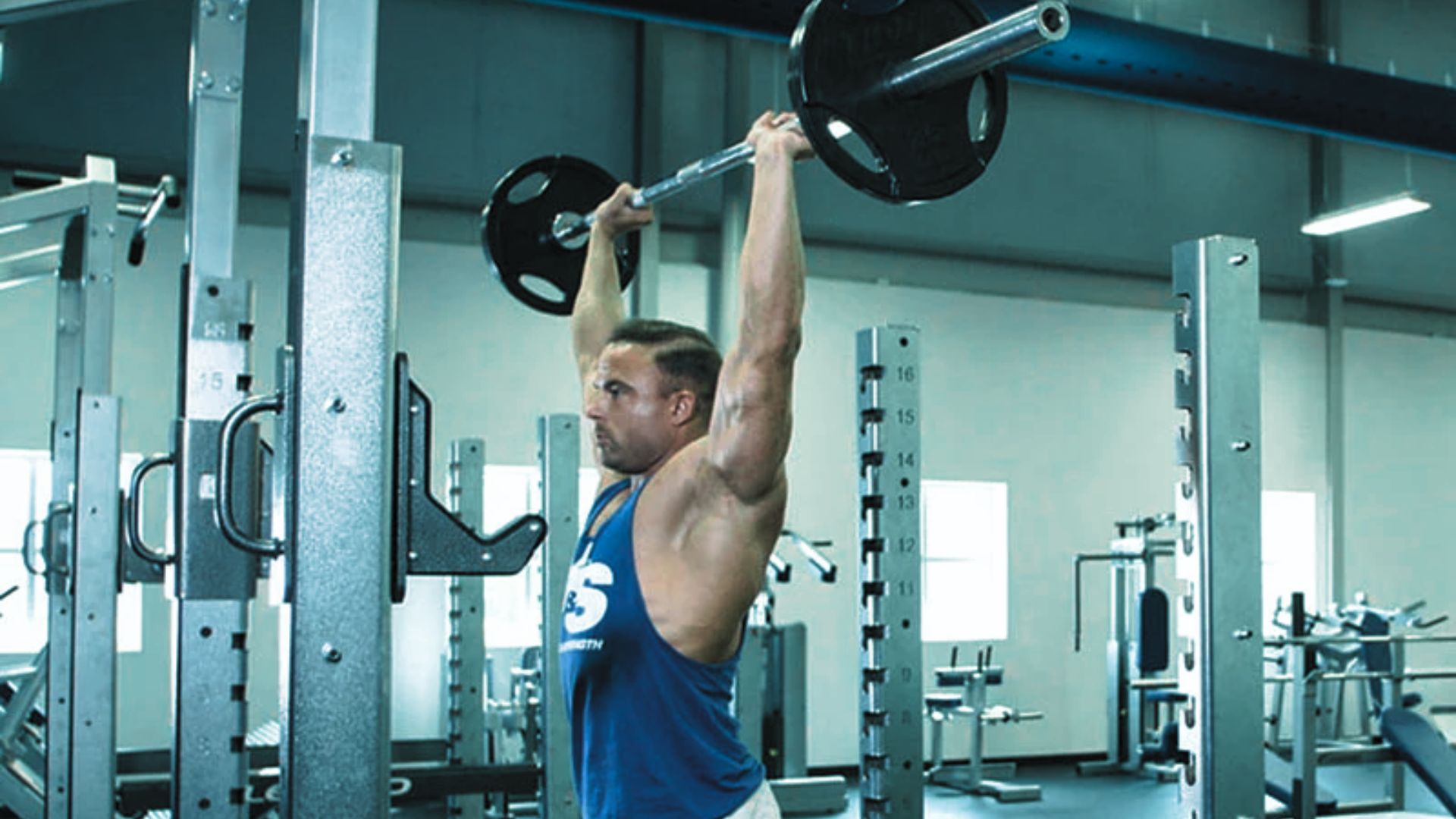A man lifts a heavy barbell overhead in a gym, showcasing strength and focus. He wears a blue tank top, surrounded by weightlifting equipment.
